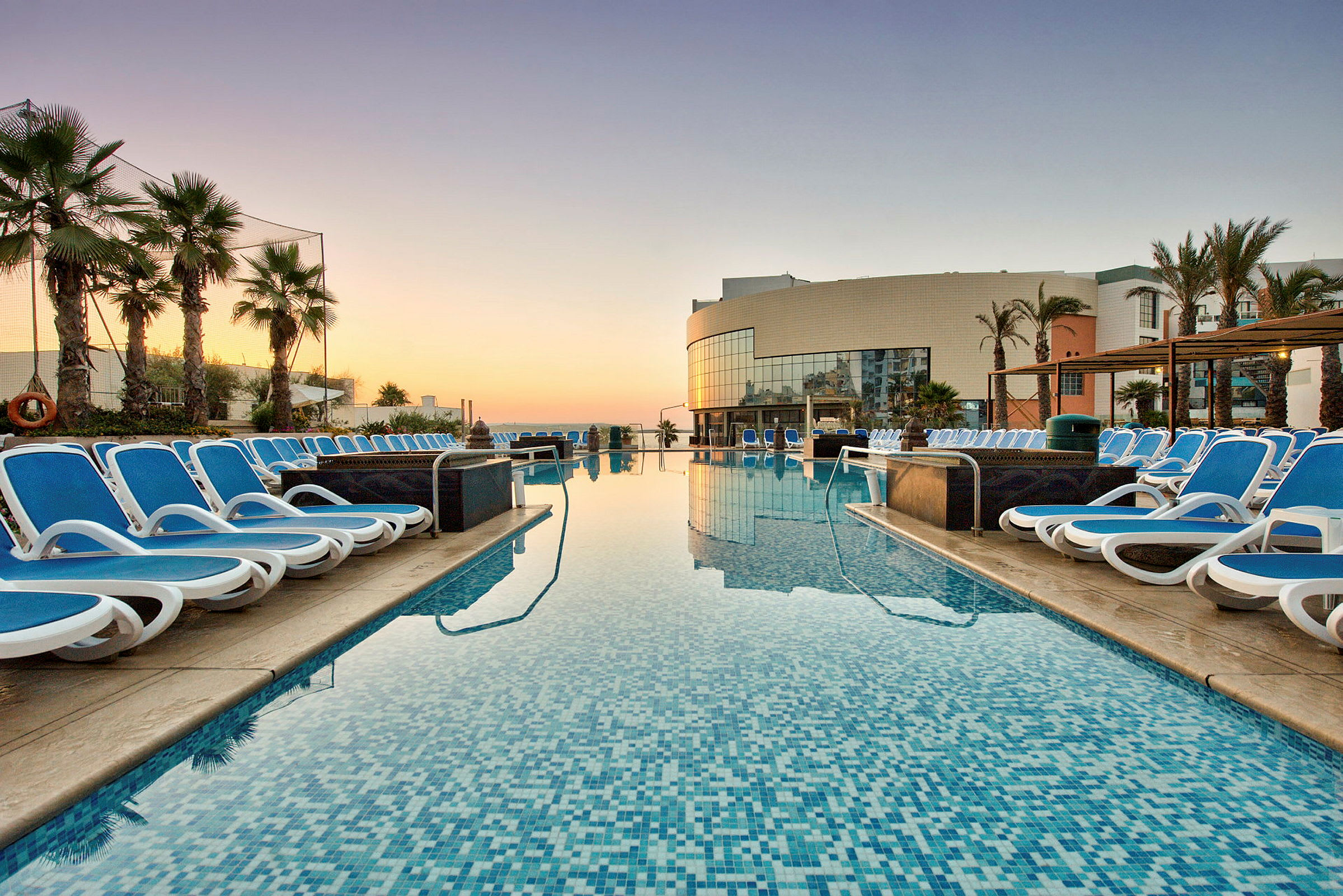 En udendørs swimmingpool omgivet af liggestole og palmer med en hotelbygning i baggrunden ved solnedgang. (AI-genereret tekst)