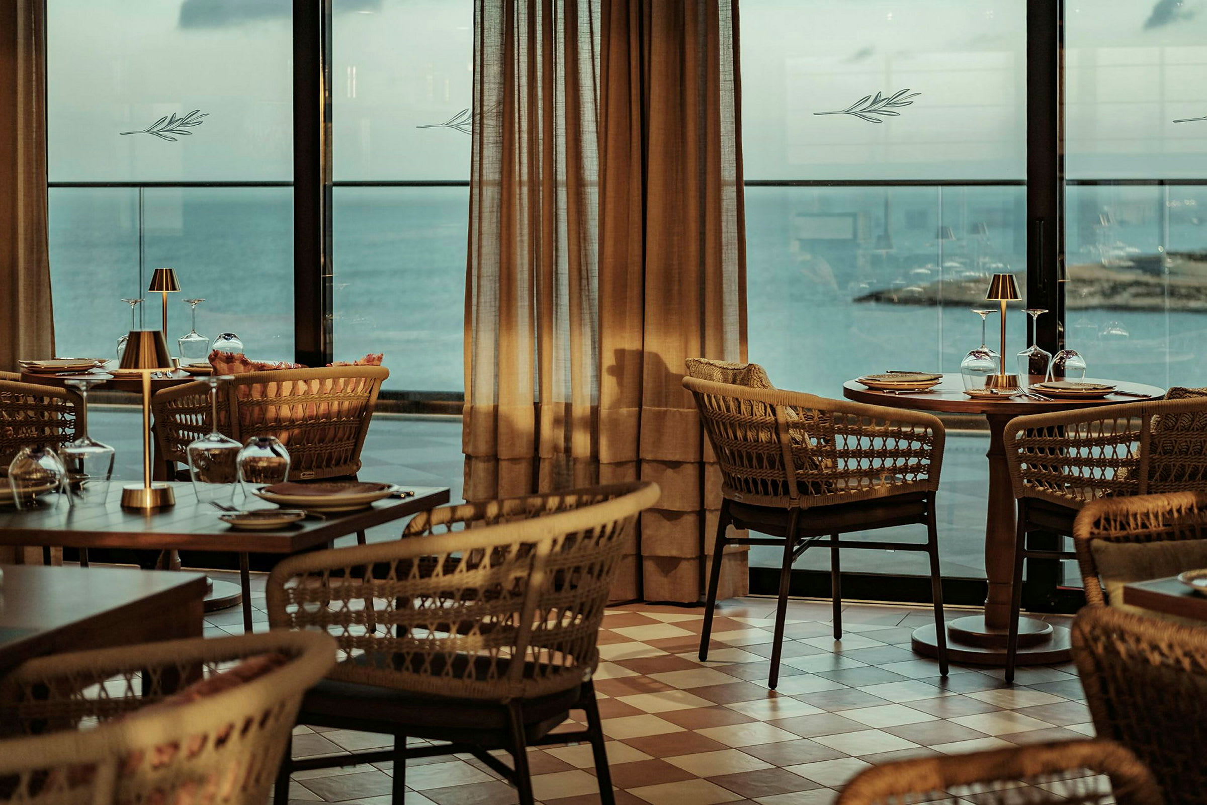 En restaurant med flettede stole og dækkede træborde med udsigt over havet gennem store vinduer. (AI-genereret tekst)