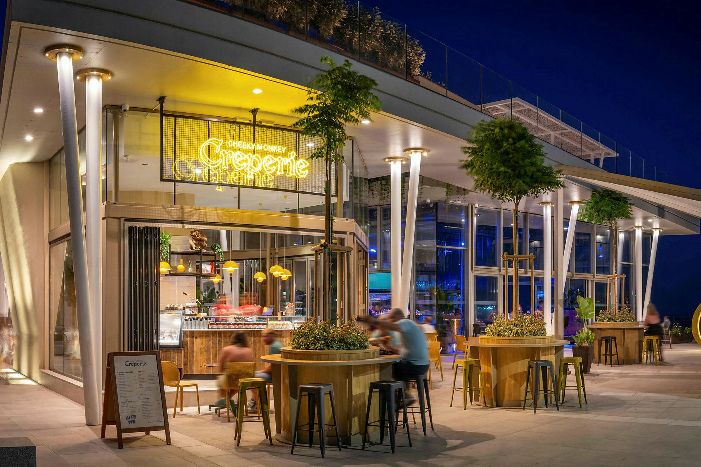 En moderne restaurant eller café med oplyst skiltning, udendørs siddepladser og en godt oplyst facade om aftenen. Træer og plantedekorationer omgiver områderne. (AI-genereret tekst)