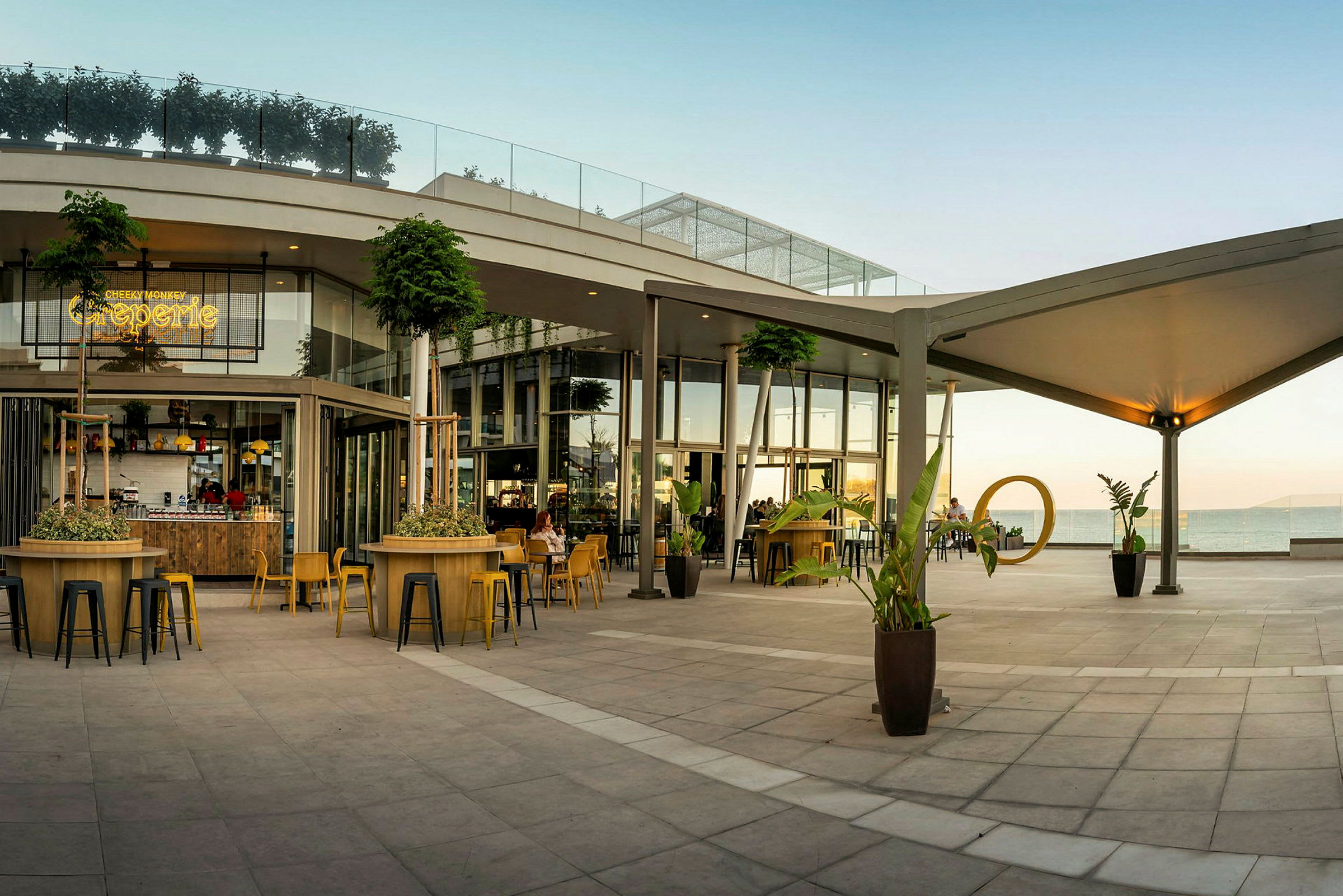 En moderne udendørs terrasse med siddepladser og beplantninger ved en cafébygning ud mod havet. (AI-genereret tekst)