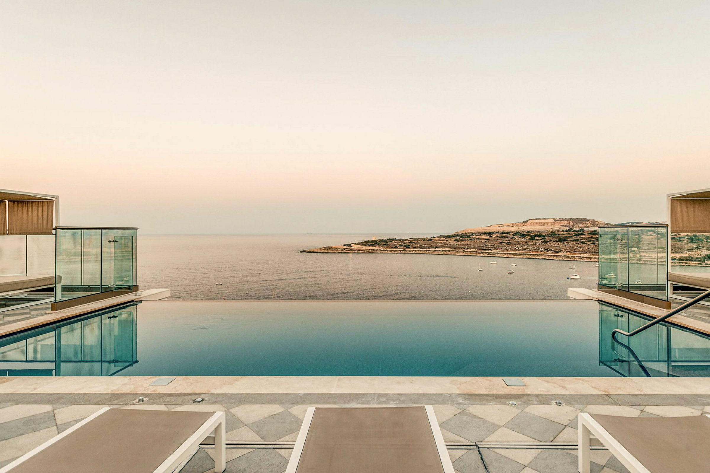 En infinitypool med udsigt over havet og en kystlinje i horisonten. Liggestole er placeret ved poolkanten. (AI-genereret tekst)