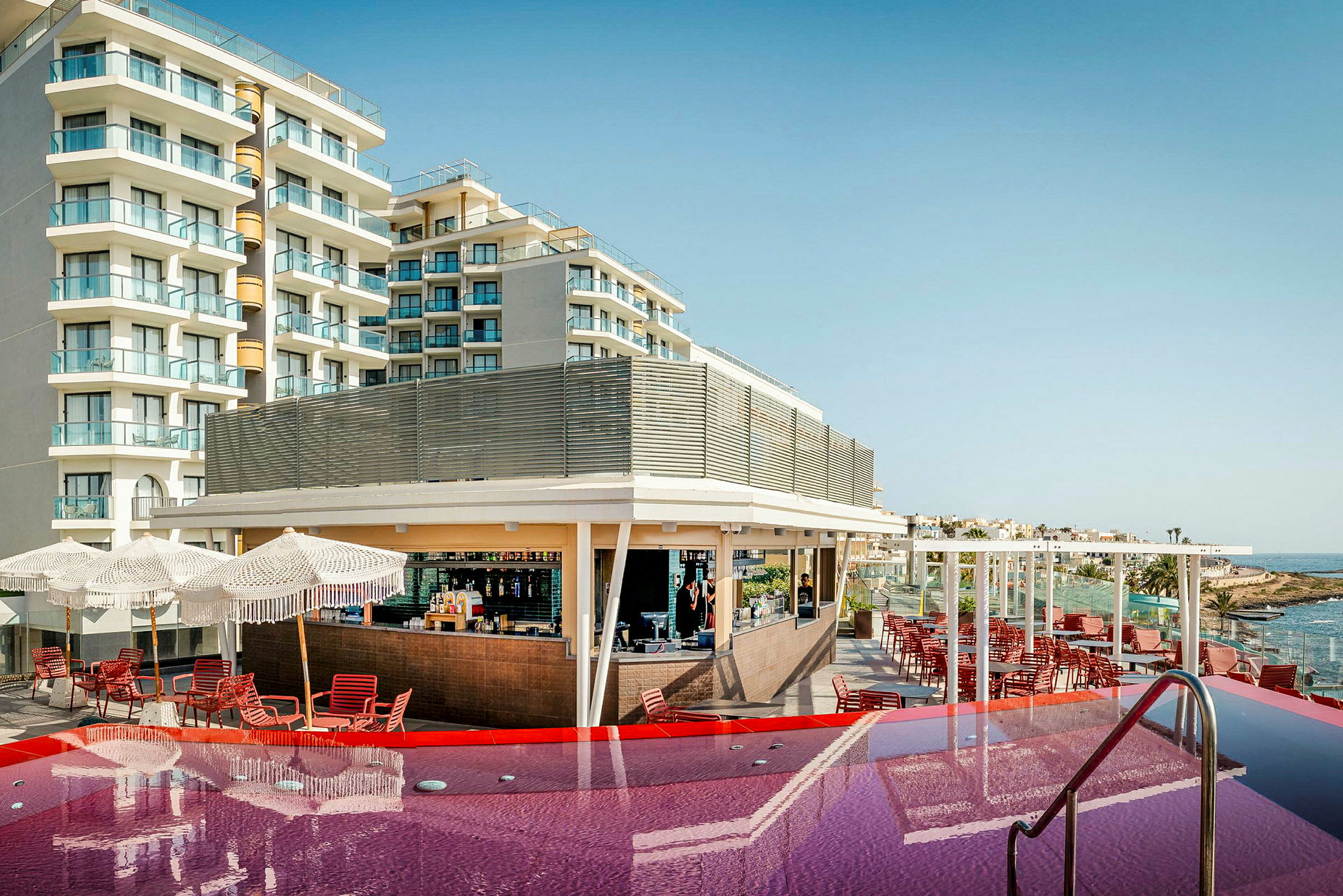 Et hotel med altaner og en pool foran, samt en bar og siddepladser under parasoller. (AI-genereret tekst)