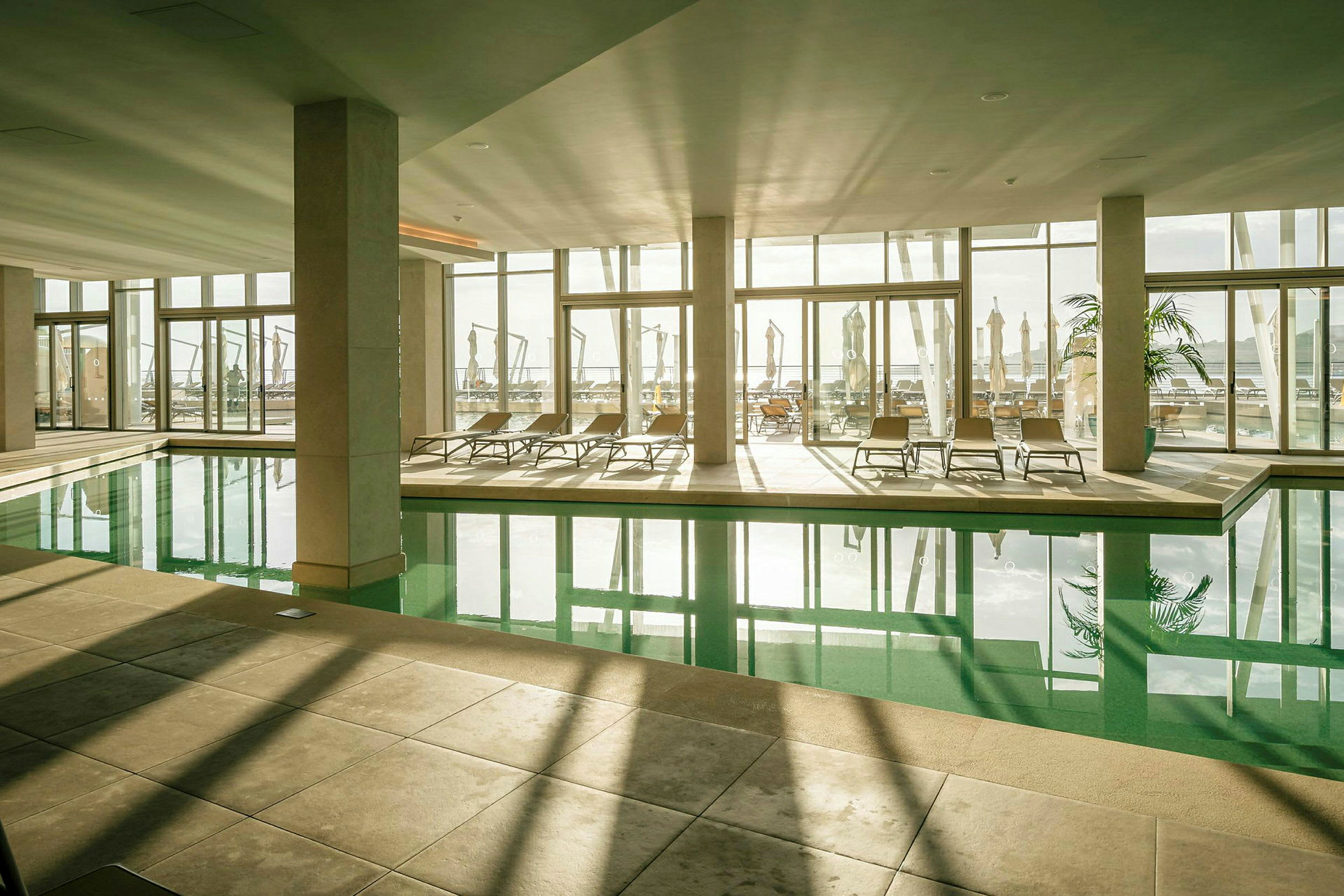 En indendørs pool i et lysfyldt rum med store vinduer mod en terrasse med liggestole. (AI-genereret tekst)