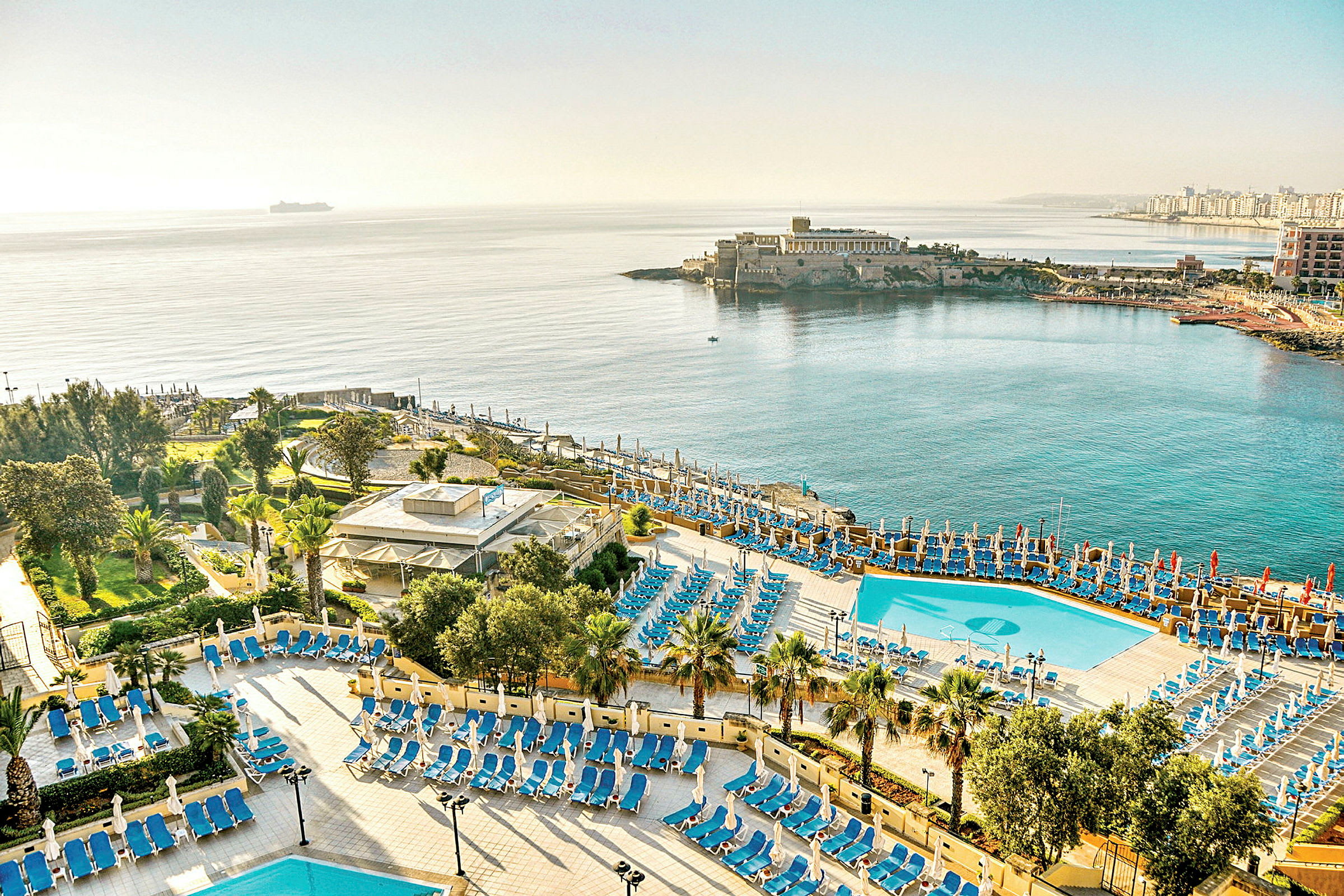 Lys og åben udsigt over et hotelområde ved havet med pools, liggestole og frodige haver. (AI-genereret tekst)