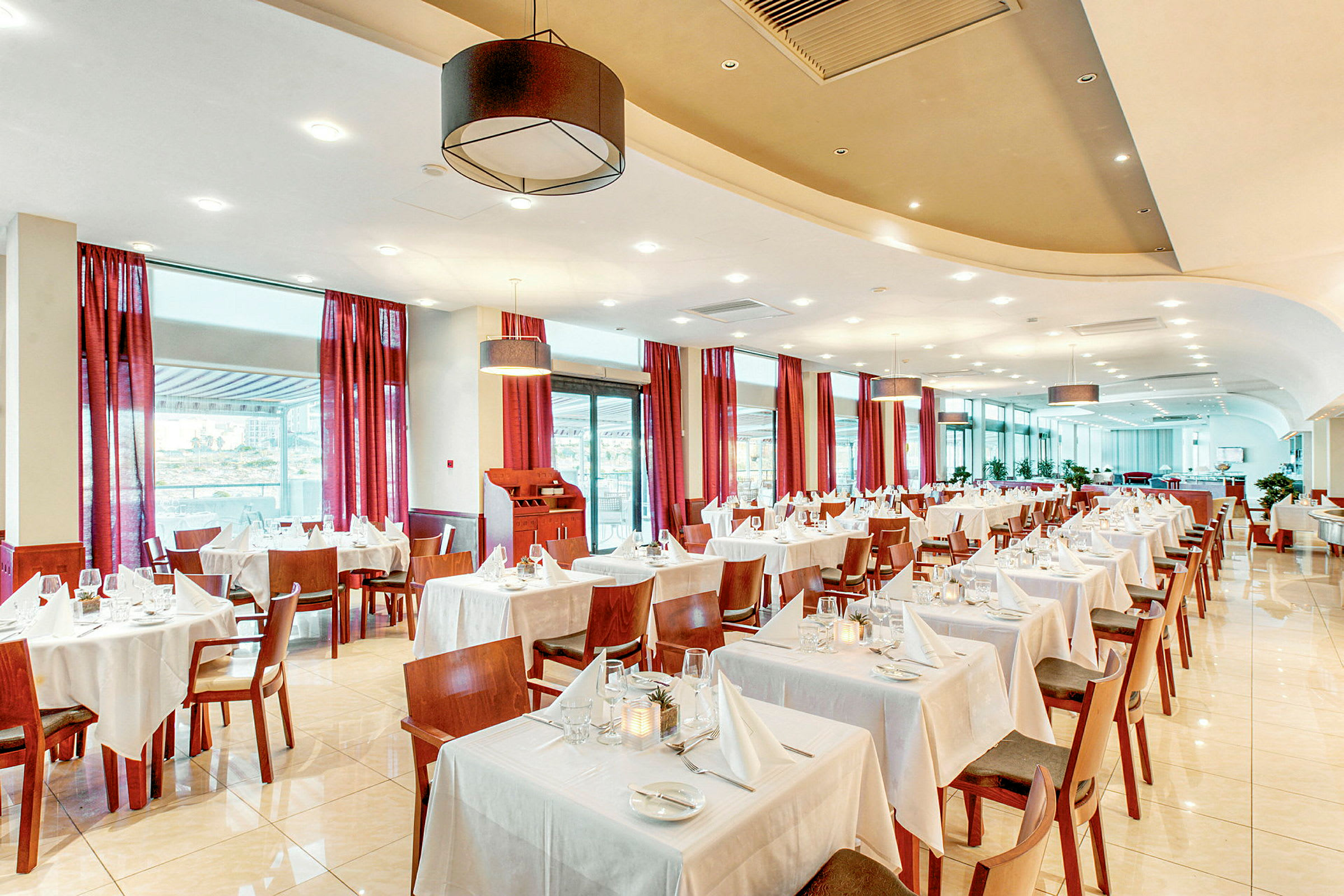 En lys restaurant med borde dækket med hvide duge og røde gardiner ved vinduerne. (AI-genereret tekst)
