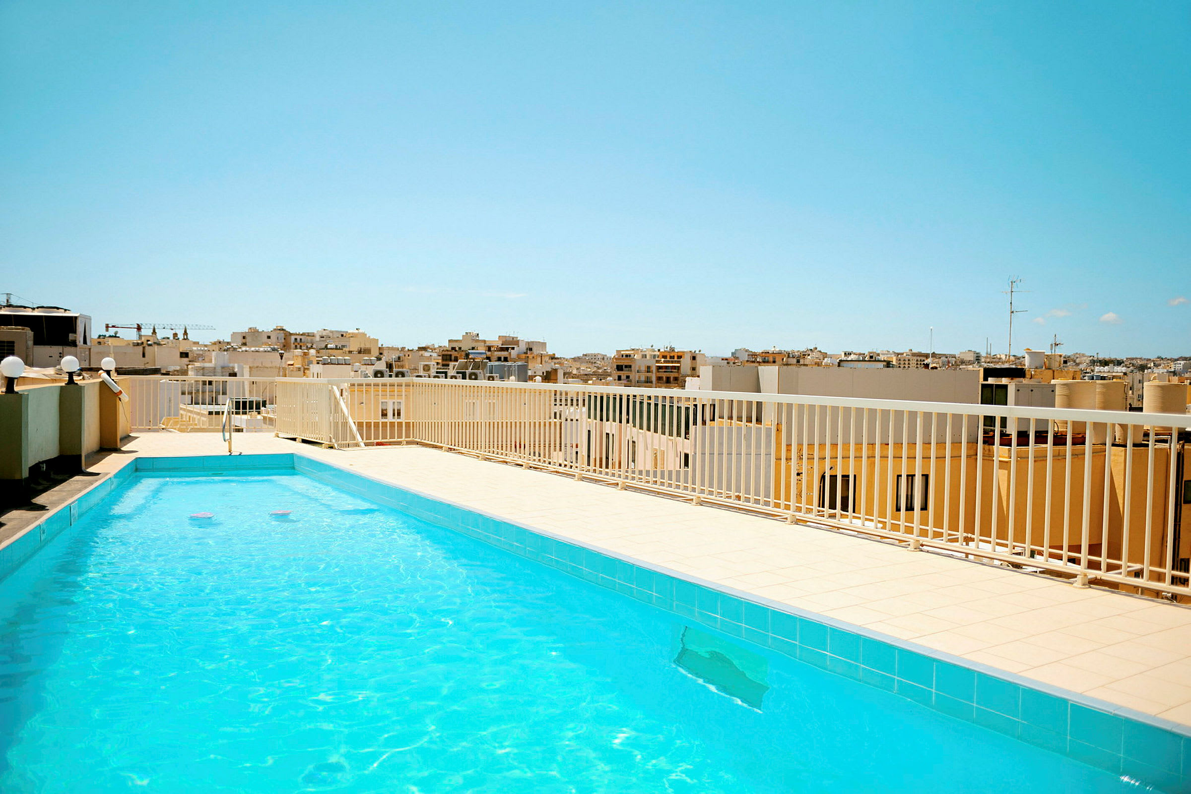 En udendørs pool med klart vand på en tagterrasse. I baggrunden ses bybebyggelse under en klar blå himmel. (AI-genereret tekst)