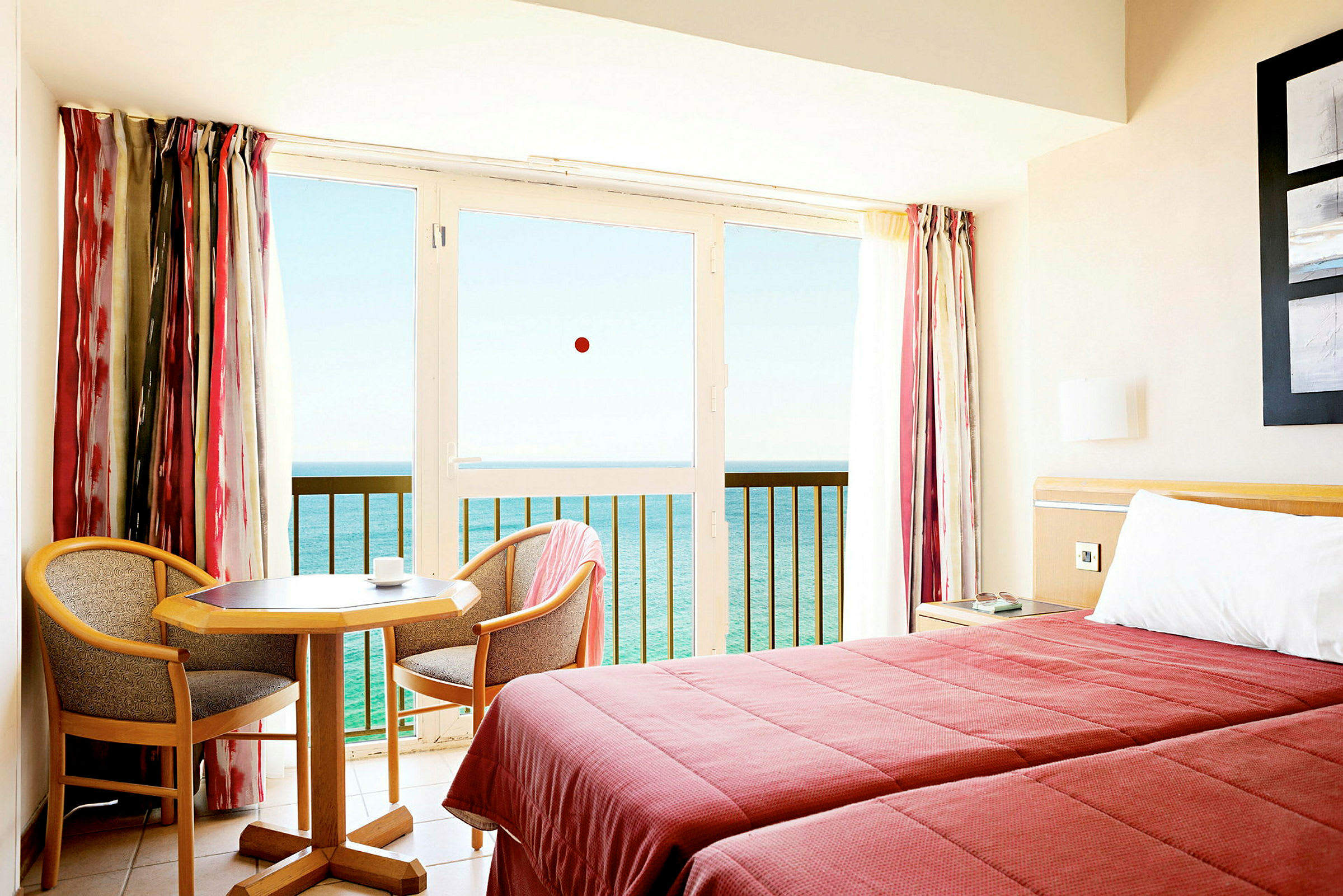 Et hotelværelse med to senge, et lille bord og to stole, samt en balkon med udsigt over havet. (AI-genereret tekst)