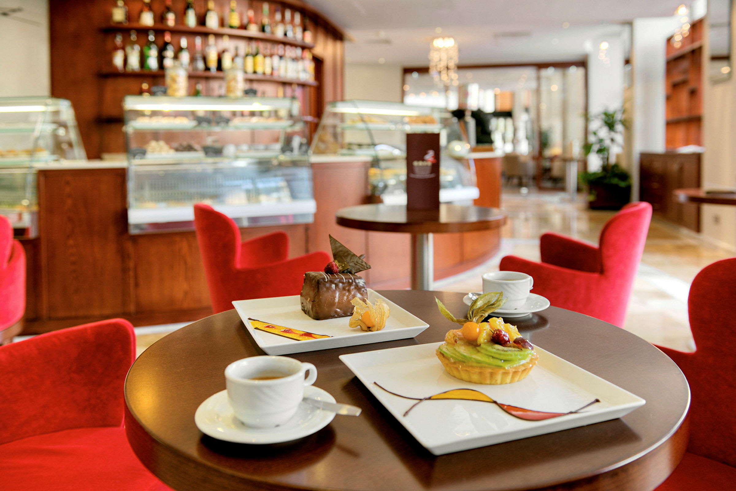 En caféinteriør med røde stole, et bord med kaffe og desserter, og en baggrund med diske og skabe. (AI-genereret tekst)