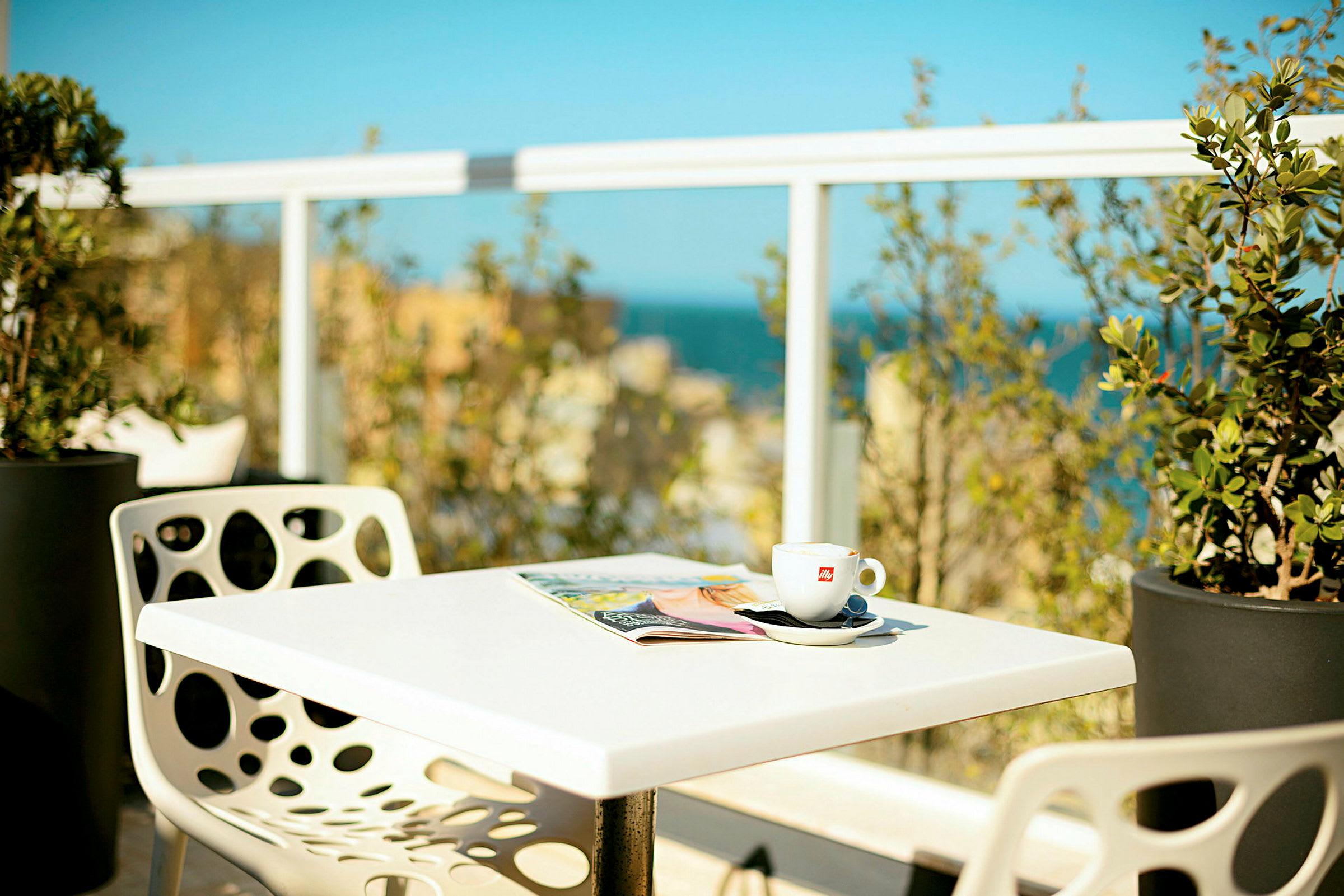 Et bord med en kaffekop og et blad, omgivet af grønne planter og med udsigt over havet i baggrunden. (AI-genereret tekst)