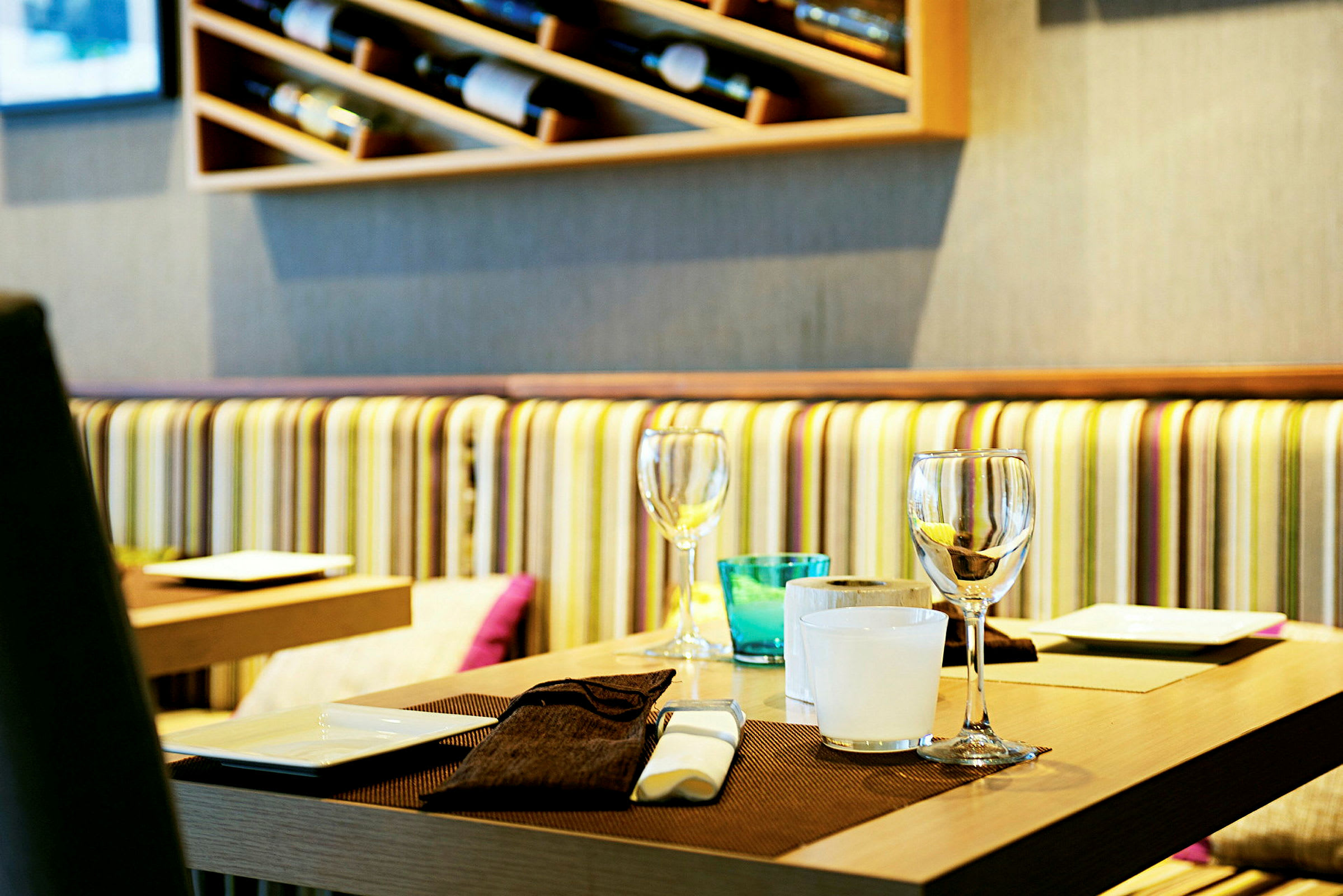 Et restaurantbord med borddækning, inklusive glas, små tallerkener og bordmåtte. Bag bordet ses en væg med hylder og stribet sofabetræk. (AI-genereret tekst)