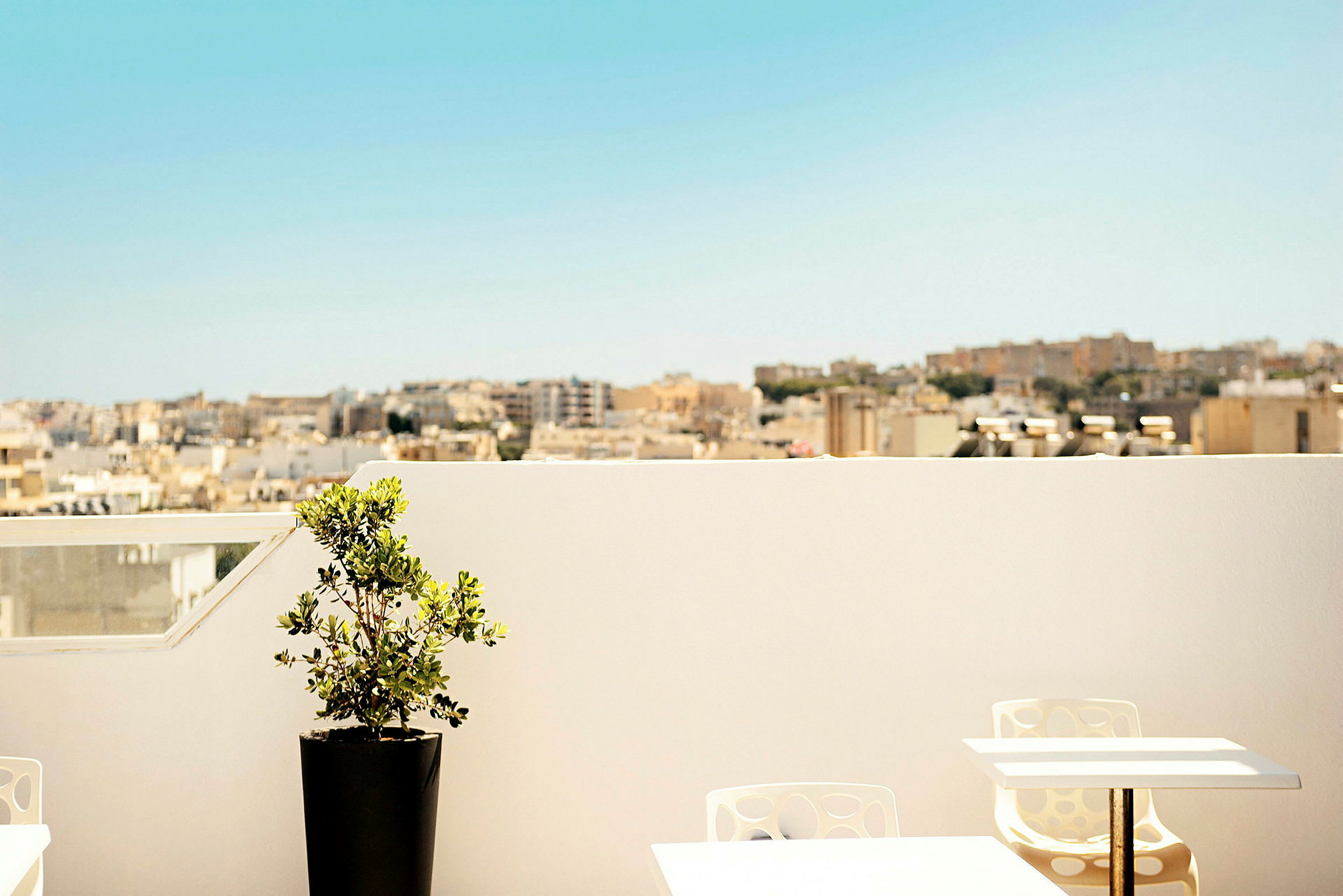 En terrasse med et bord, stole og en potteplante. I baggrunden ses en byudsigt under en klar blå himmel. (AI-genereret tekst)
