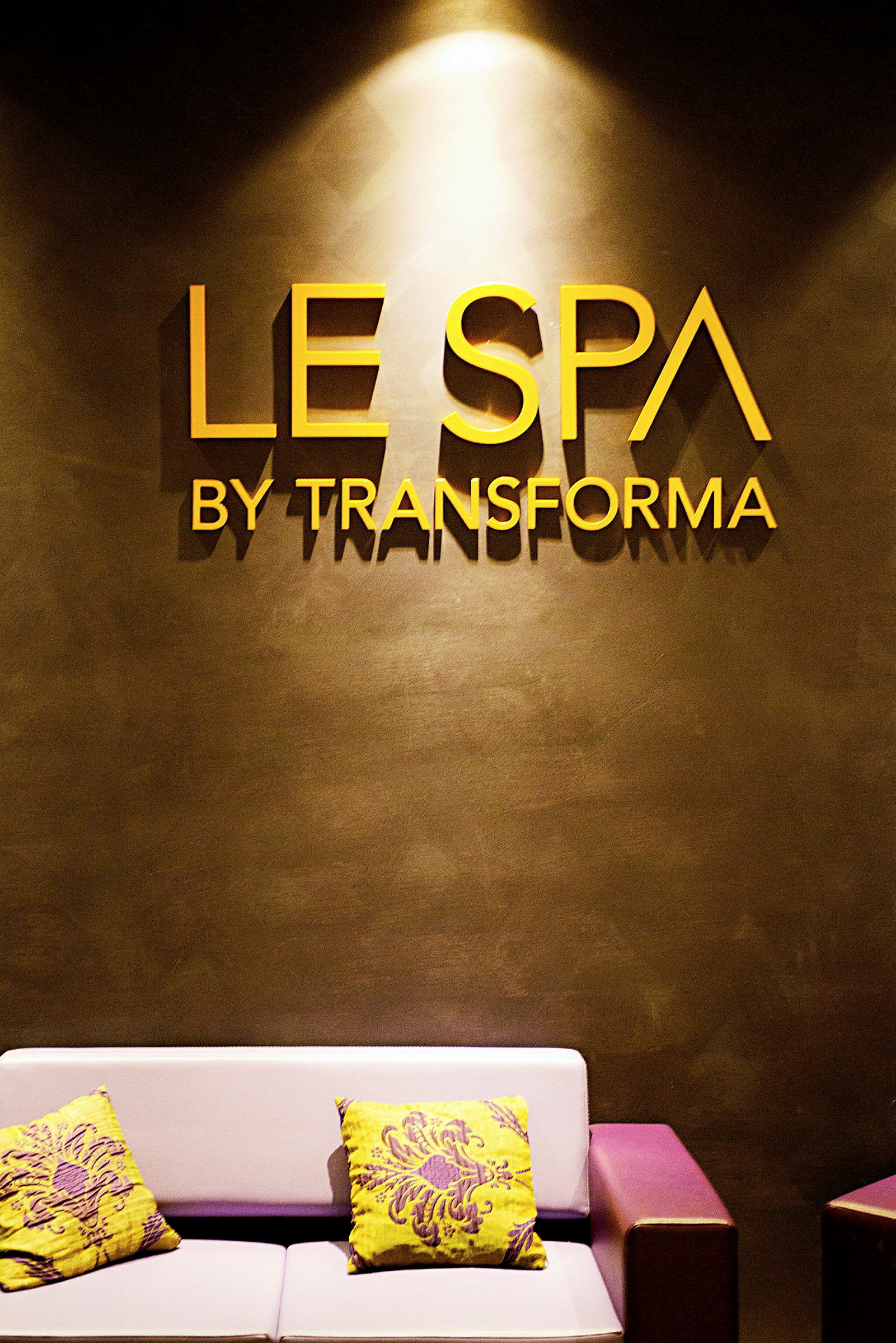 Billede af en spa-indgang med teksten "Le Spa by Transforma" på væggen. En sofa med farverige pyntepuder er synlig. (AI-genereret tekst)