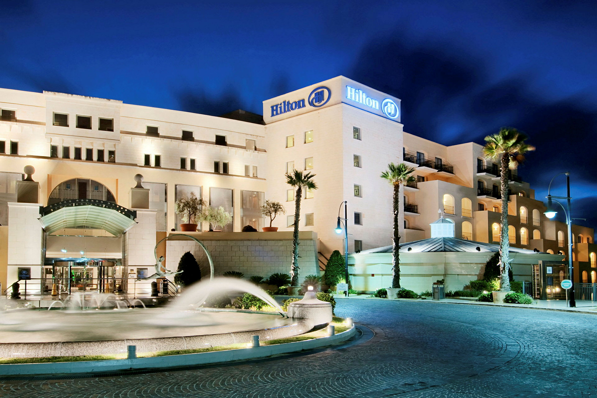 Et hotel med Hilton-logoen på facaden, oplyst under en nattehimmel. Foran er der en springvand, nogle palmer og parkering. (AI-genereret tekst)
