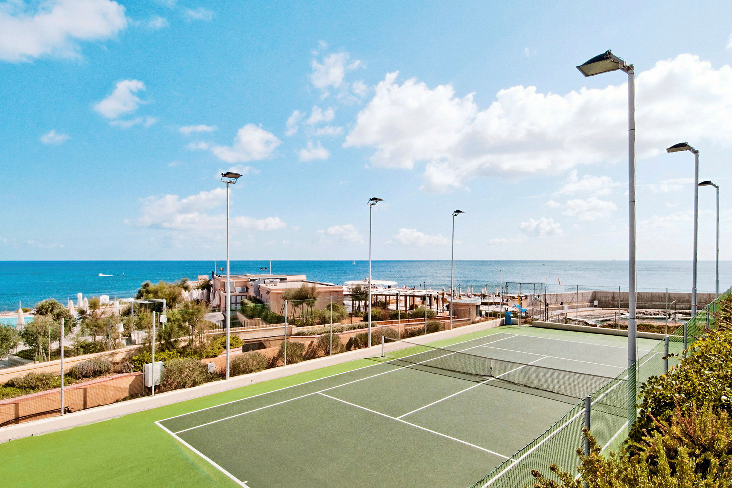 En tennisbane med grønt underlag, omgivet af hegn og lygtepæle, med havet og bygninger i baggrunden. (AI-genereret tekst)
