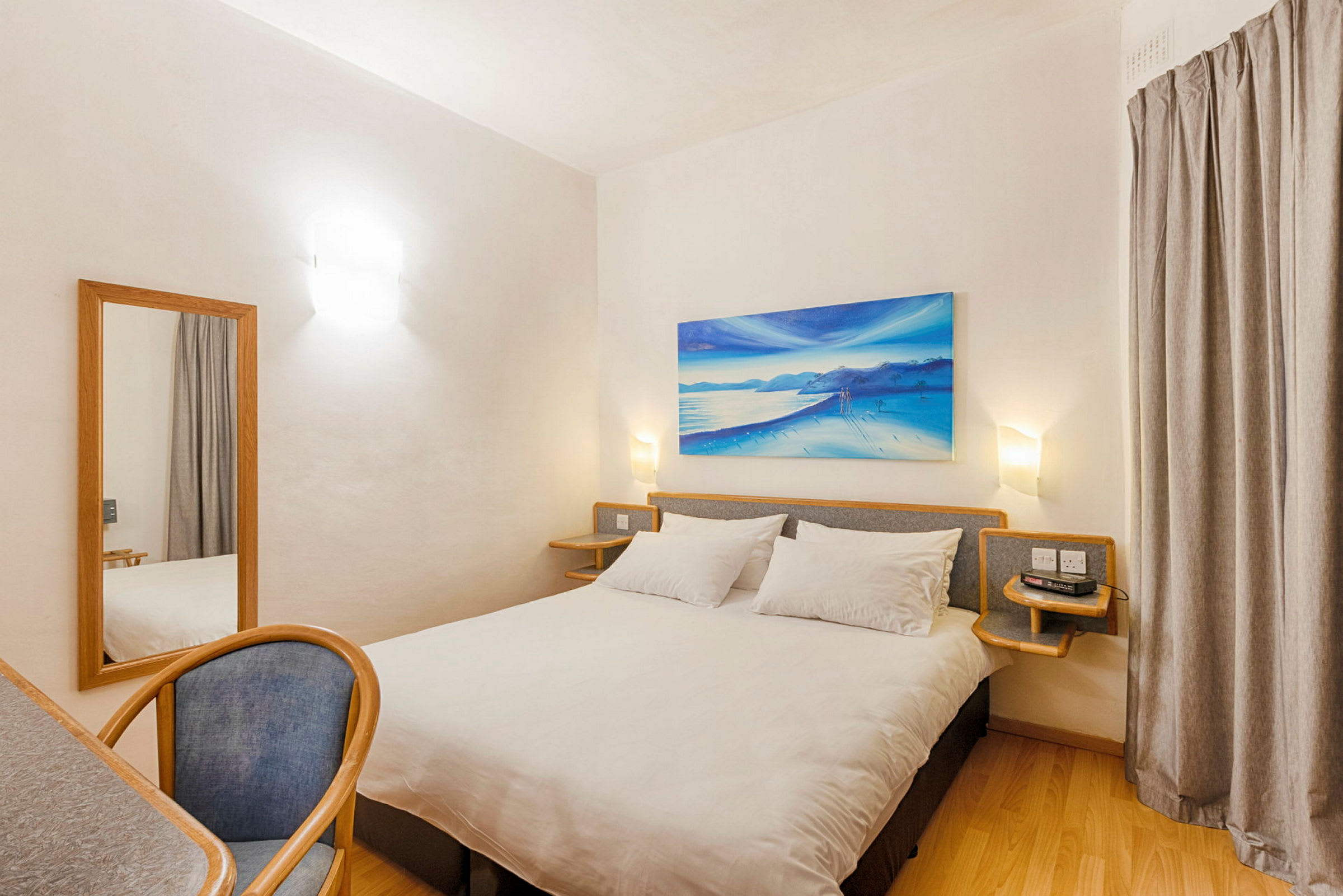 Et hotelværelse med en dobbeltseng, et skrivebord med en stol og et spejl. Værelset har lyse vægge og en vægdekoration med strandmotiv. (AI-genereret tekst)