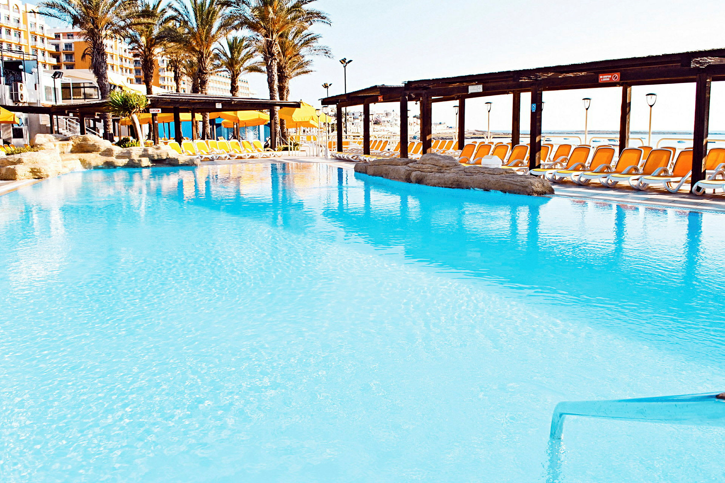 En udendørs swimmingpool med klart blåt vand, omgivet af liggestole med orange hynder og parasoller. Palmer og hotelbygninger ses i baggrunden. (AI-genereret tekst)