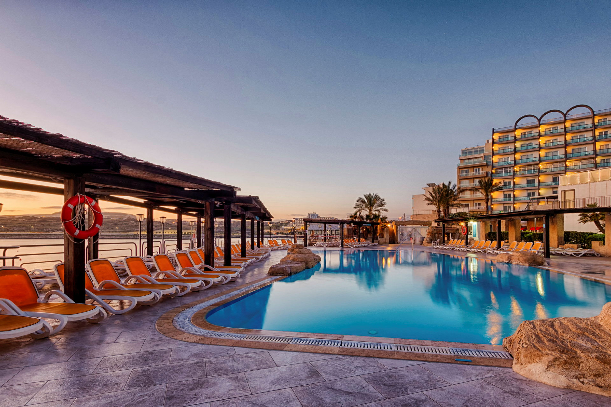 Billede af et hotelpoolområde med liggestole, parasoller og en stor swimmingpool i solnedgangen. (AI-genereret tekst)
