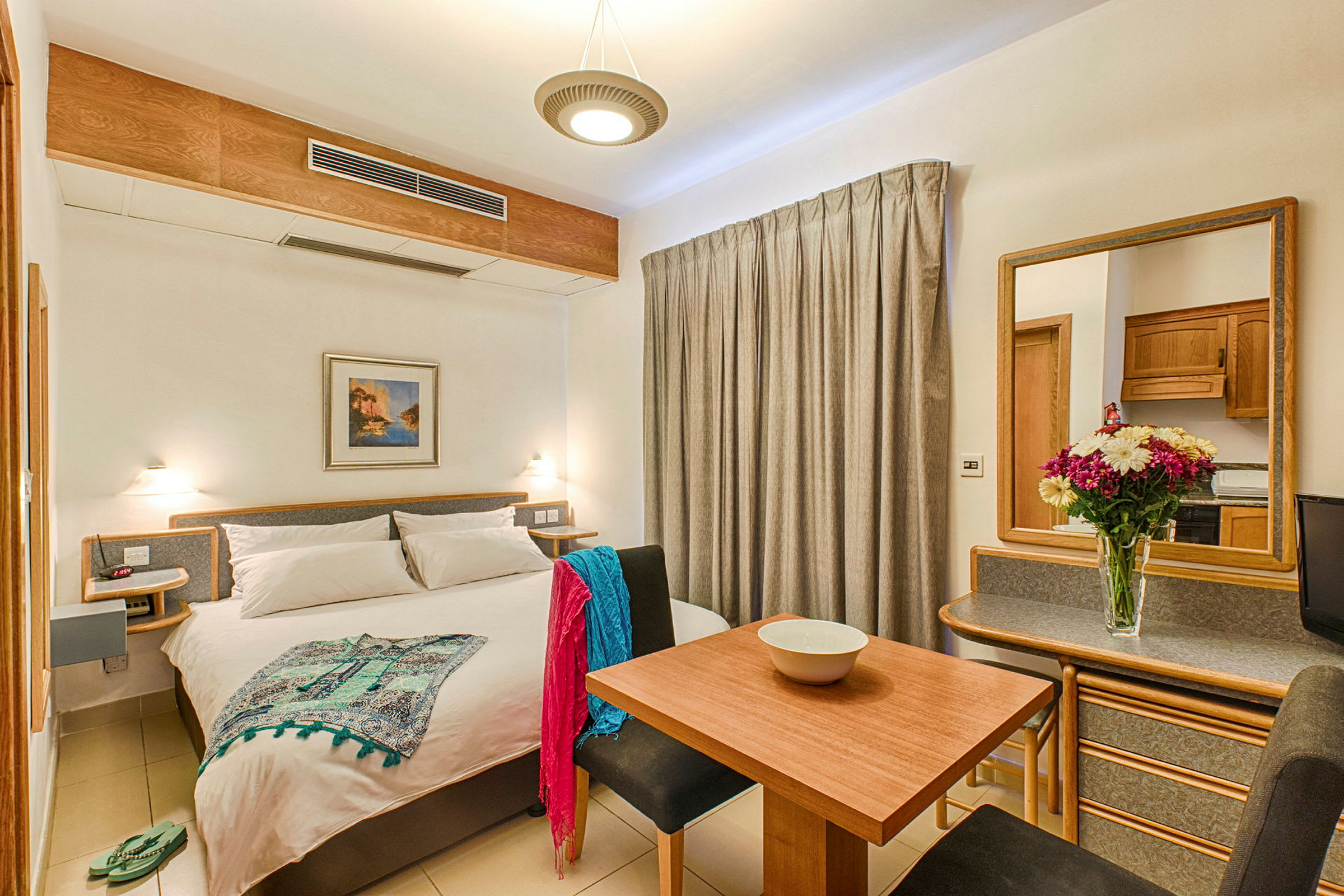 Et lyst hotelværelse med en dobbeltseng, spisebord, siddepladser og et spejl. Dekoreret med en blomsterkrukke på bordet. (AI-genereret tekst)