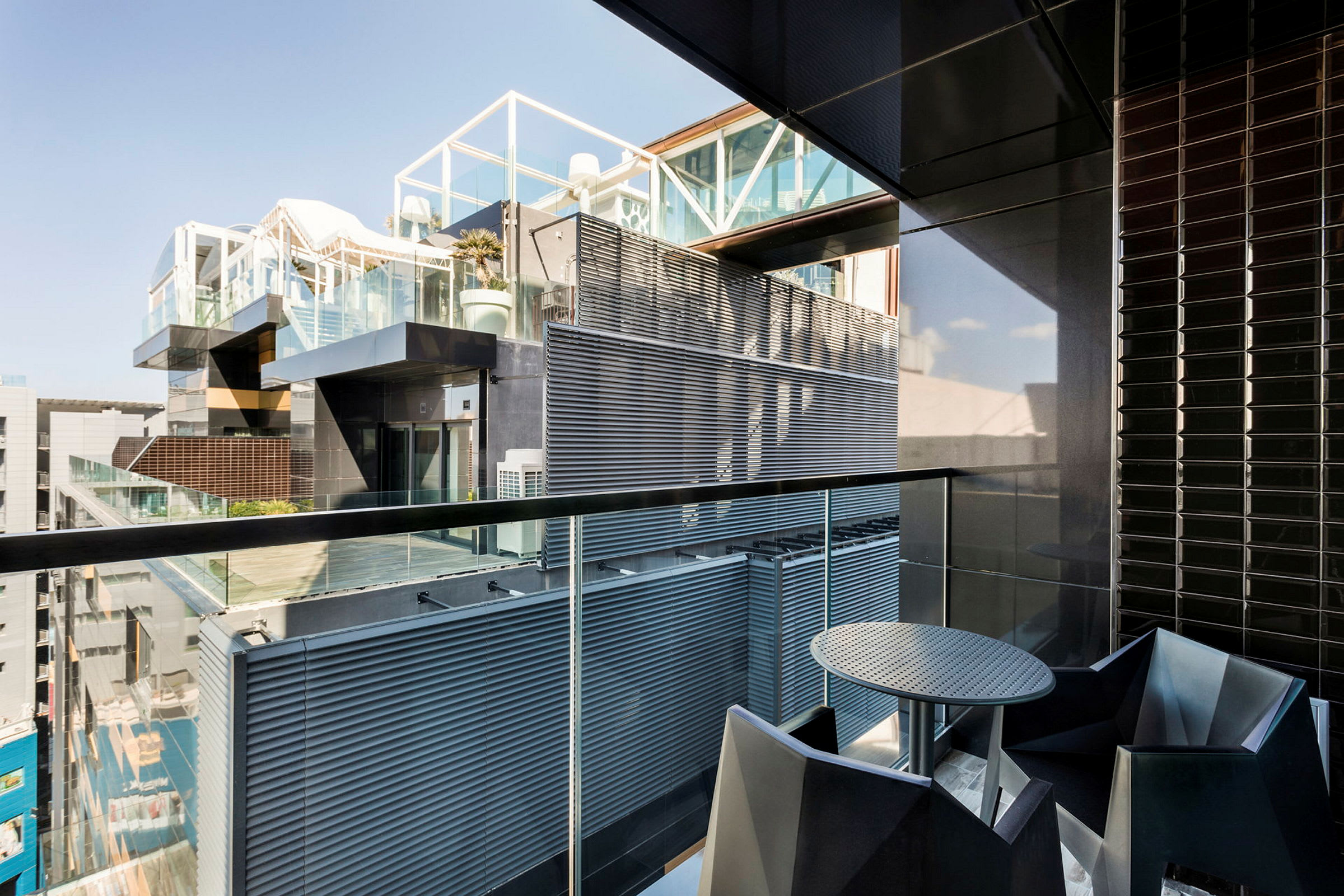 En balkon med moderne indretning, inklusive et rundt bord og stole, med udsigt over en bygning med glas- og metalfacader. (AI-genereret tekst)