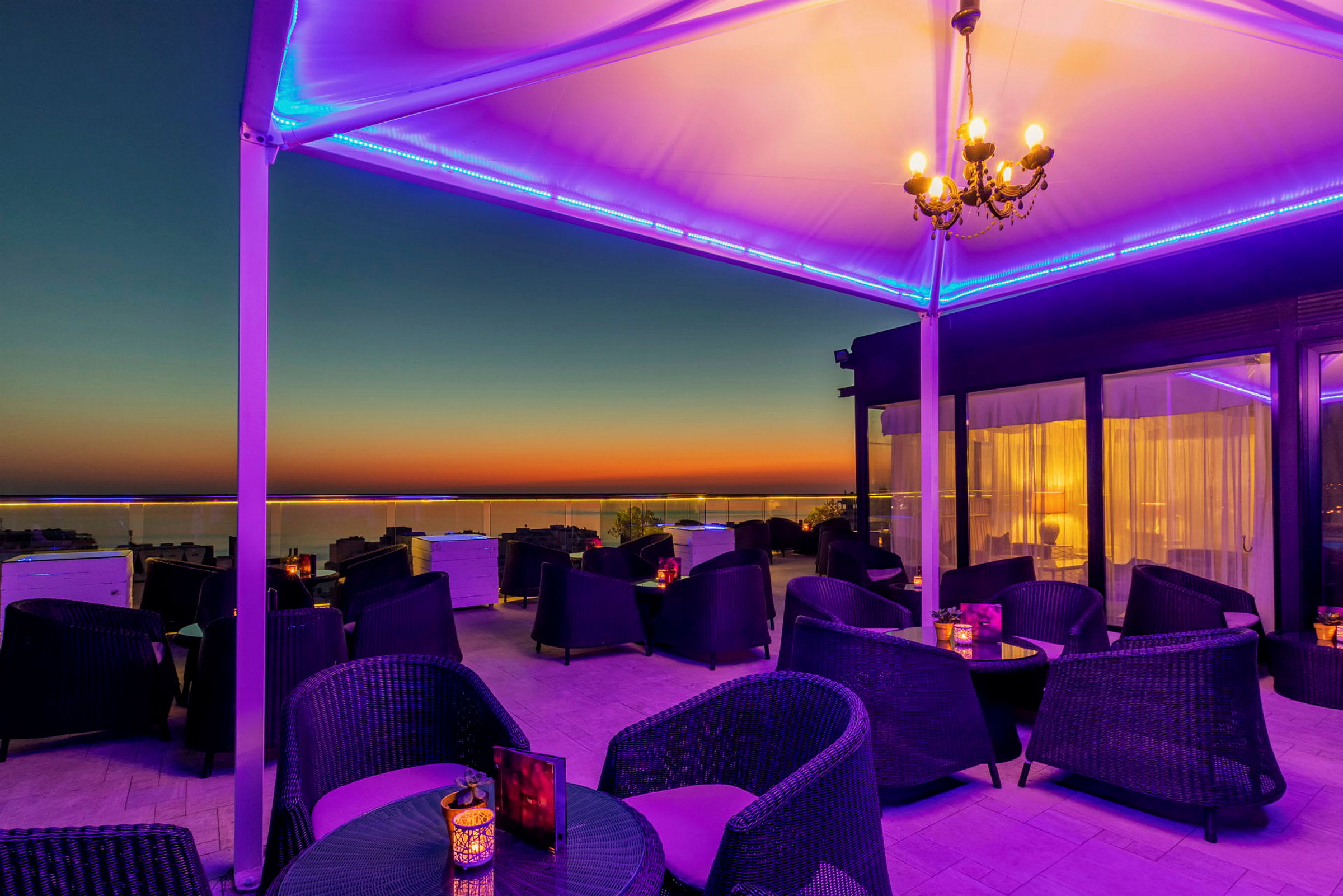 En tagterrasse med siddepladser og borde, oplyst med lilla og gule farver mod en solnedgang. (AI-genereret tekst)