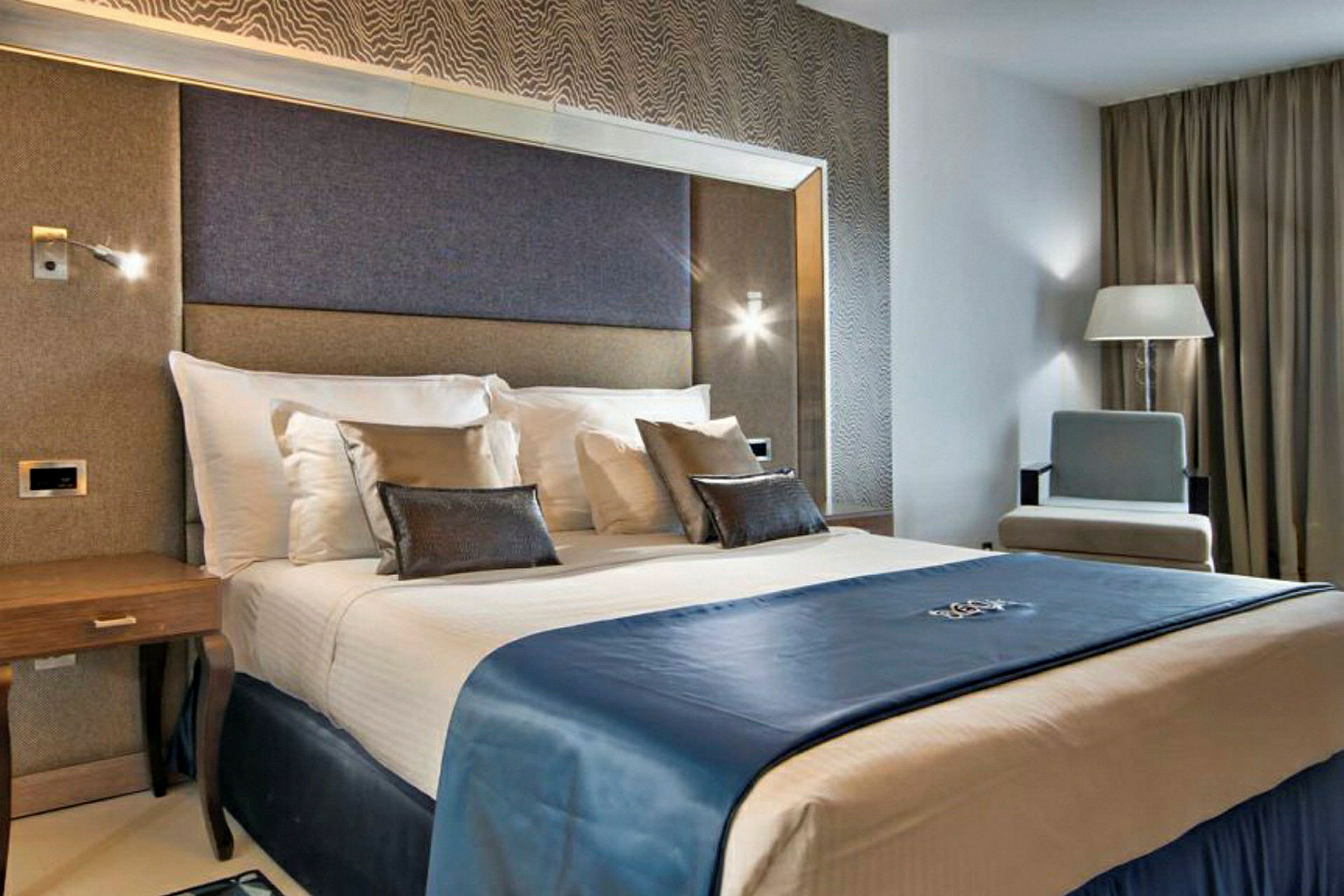 Et hotelværelse med moderne interiør, der viser en dobbeltseng med hvidt sengetøj og et blåt sengetæppe, omgivet af elegante møbler. (AI-genereret tekst)