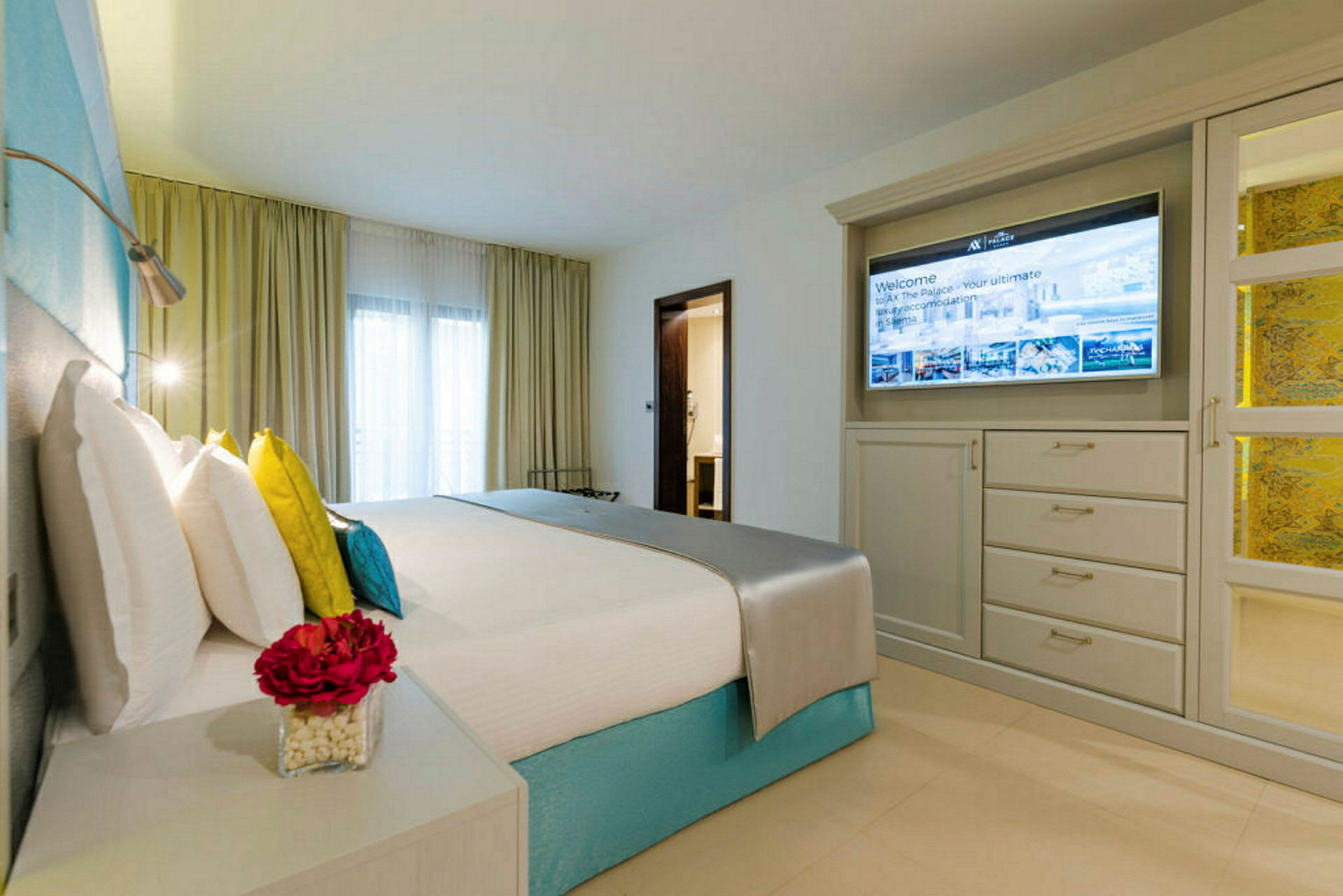 Et moderne hotelværelse med en opredt seng, farverige puder, et natbord med en blomstervase og en væg med tv foran et skab. (AI-genereret tekst)