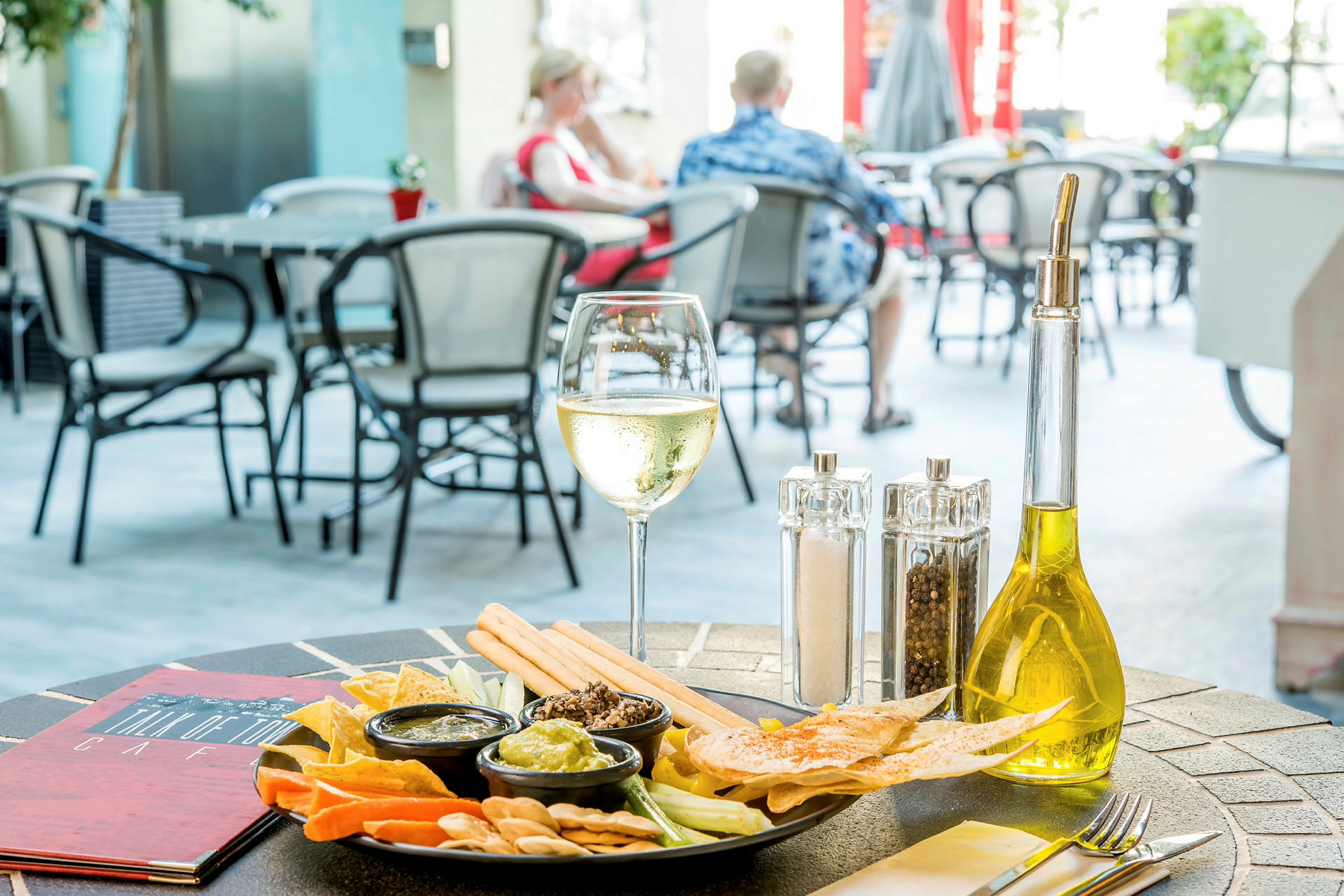 Et bord på en udendørsrestaurant med en tallerken med snacks, små skåle med dip, et glas hvidvin og en flaske olivenolie. (AI-genereret tekst)