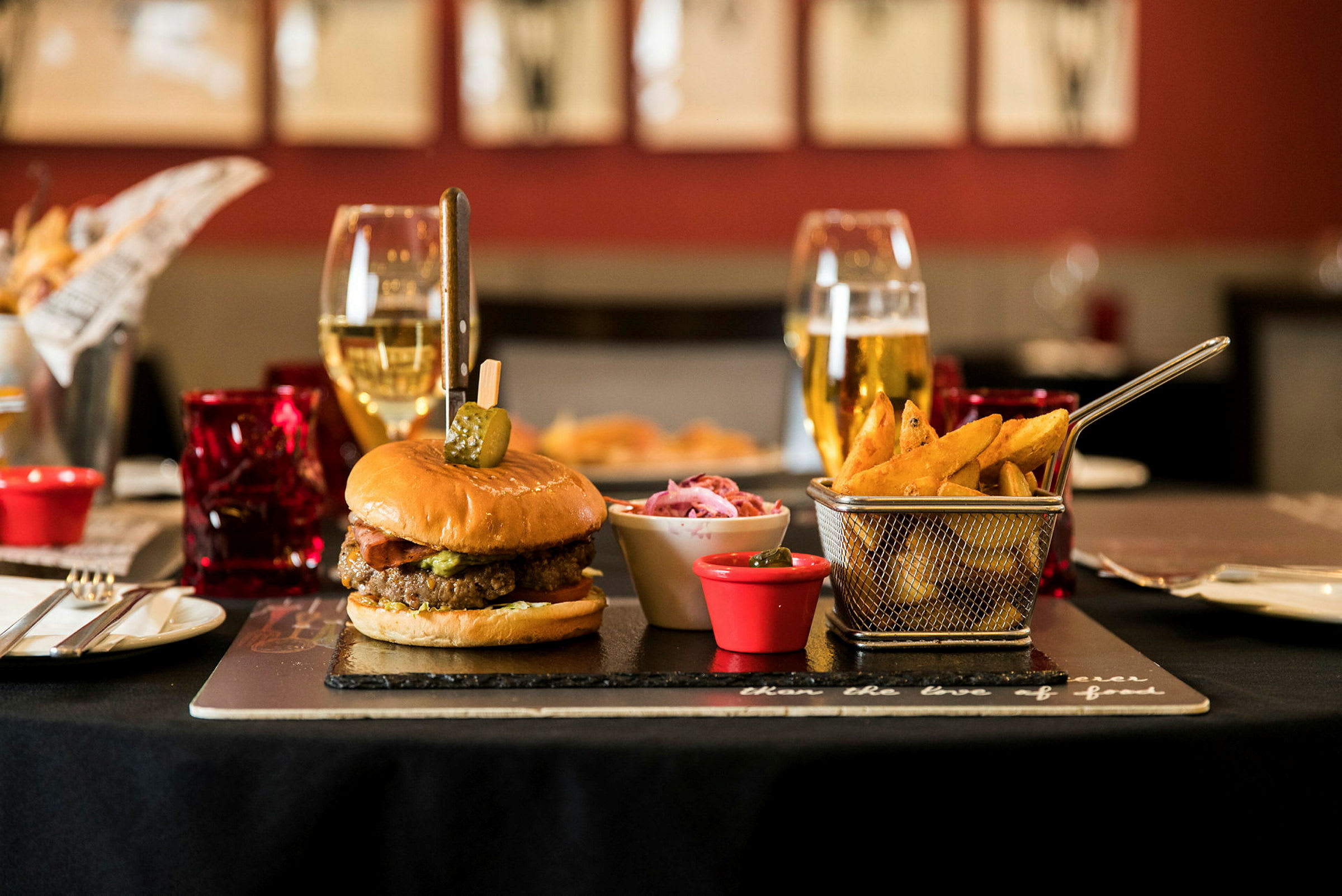 En burger med pommes frites og tilbehør på et bord, omgivet af glas og bestik. (AI-genereret tekst)