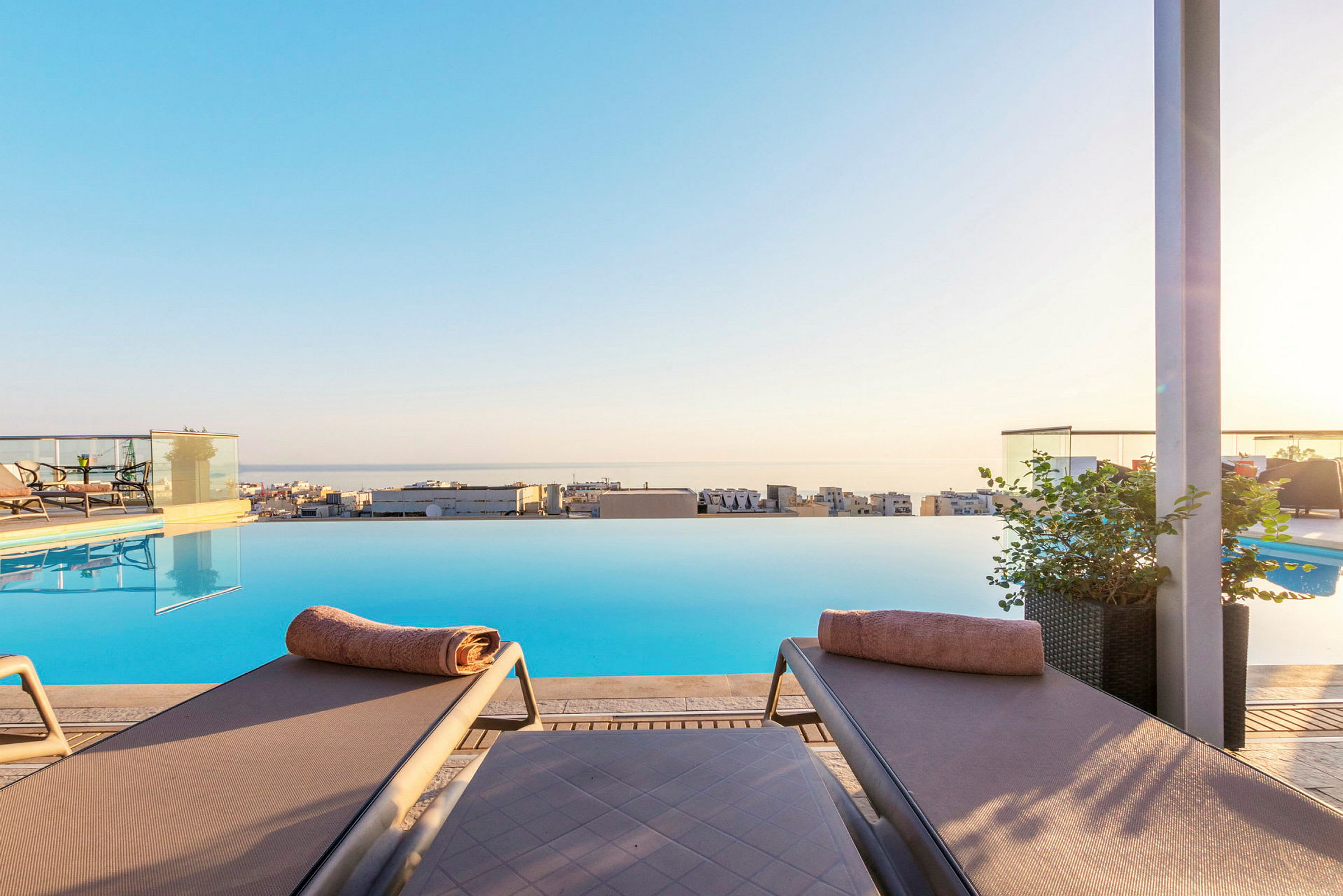 To solsenge med håndklæder ved en infinity-pool med udsigt over byen og havet. (AI-genereret tekst)