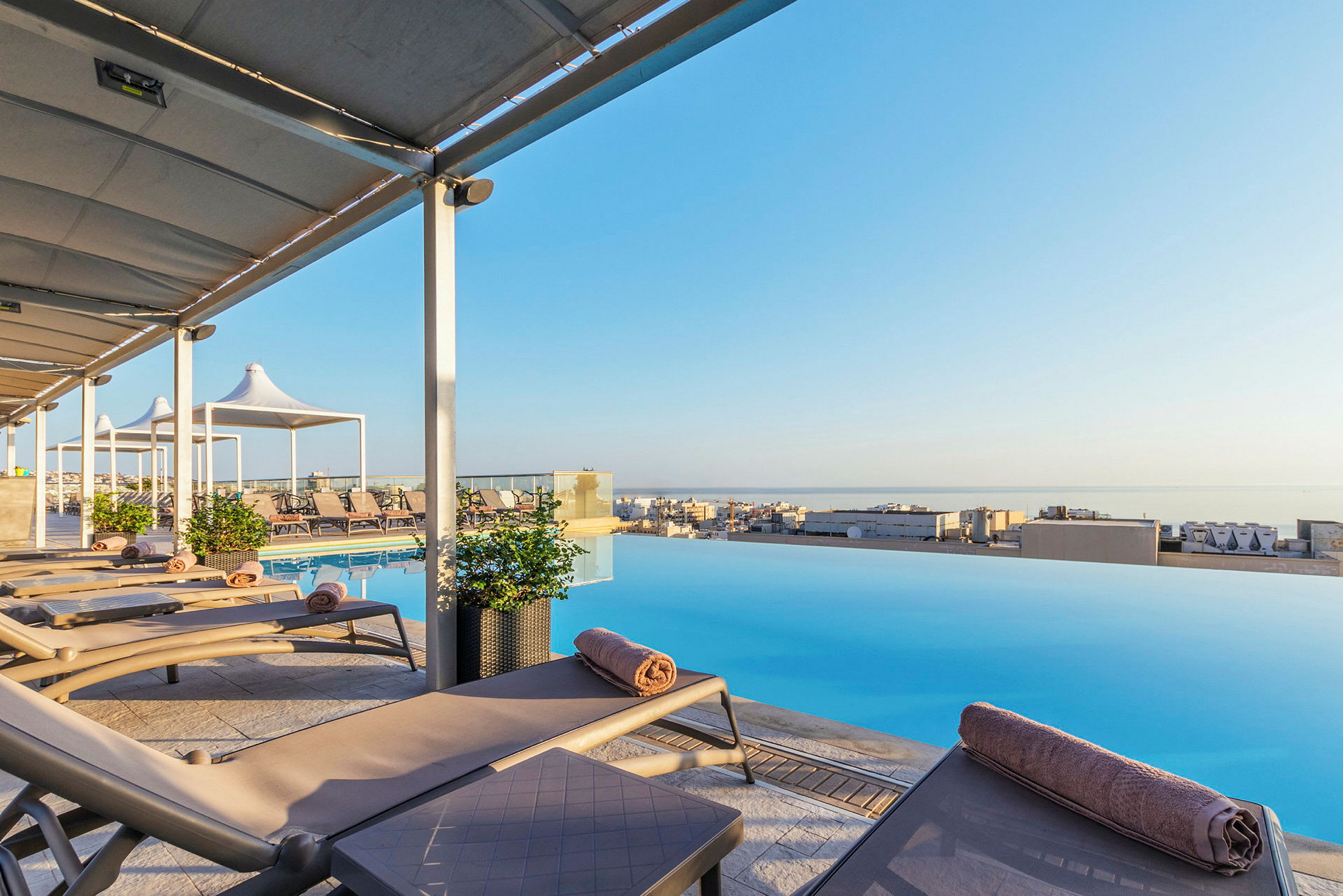 Liggestole ved en infinity-pool med udsigt over en by og havet. (AI-genereret tekst)