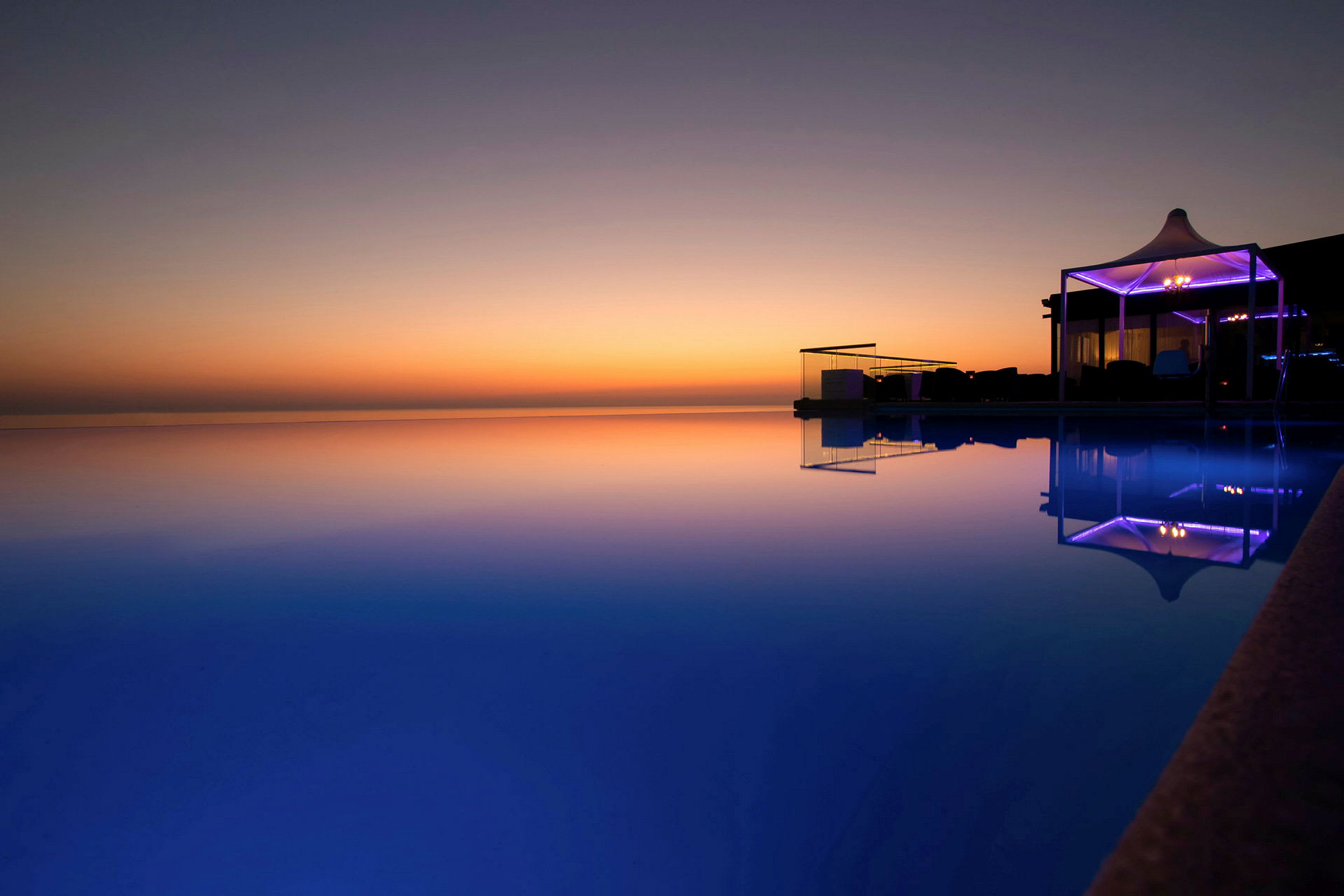 En infinitypool med udsigt over en solnedgang. Poolen reflekterer den farverige himmel og et oplyst område ved kanten. (AI-genereret tekst)