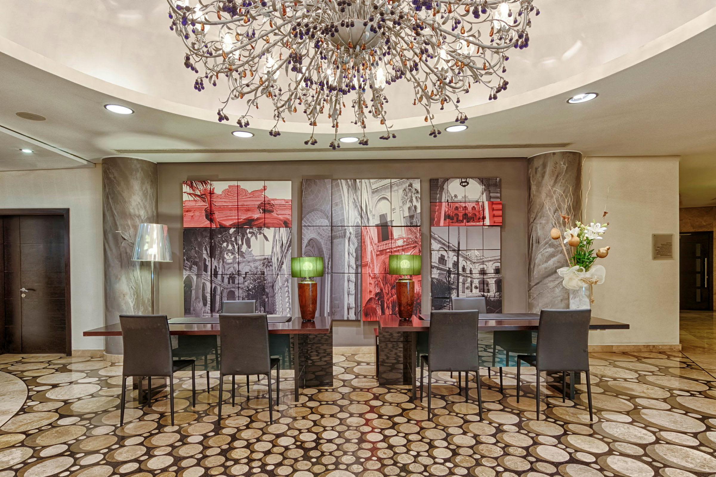 En elegant hotellobby med en stor dekorativ loftslampe, der hænger over flere skriveborde med stole, malerier på væggen bagved og smukke dekorationer som blomster og bordlamper. (AI-genereret tekst)