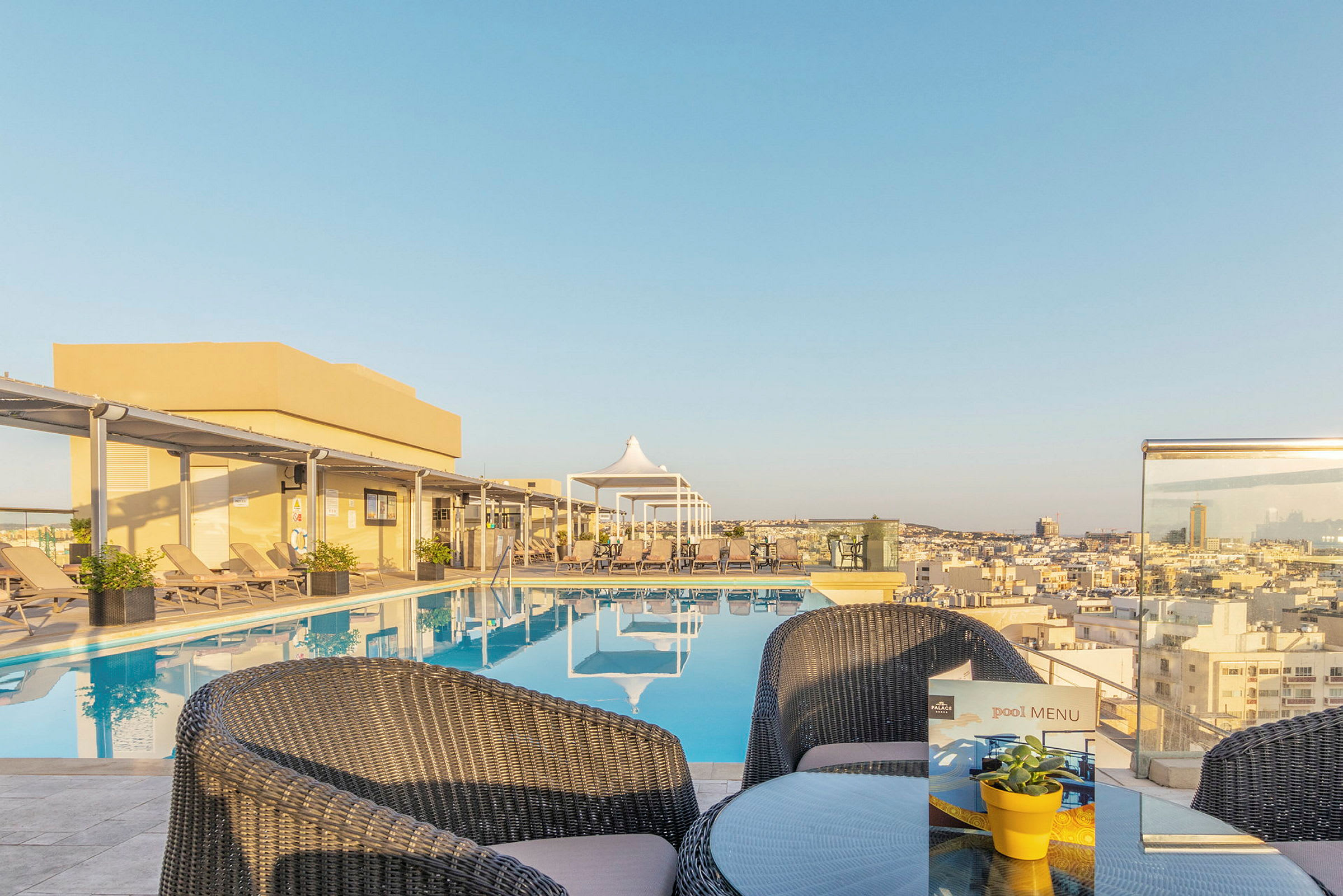 Billede af en swimmingpool på en hotelterrasse med udsigt over byen, omgivet af solsenge og et mindre bord med stole. (AI-genereret tekst)