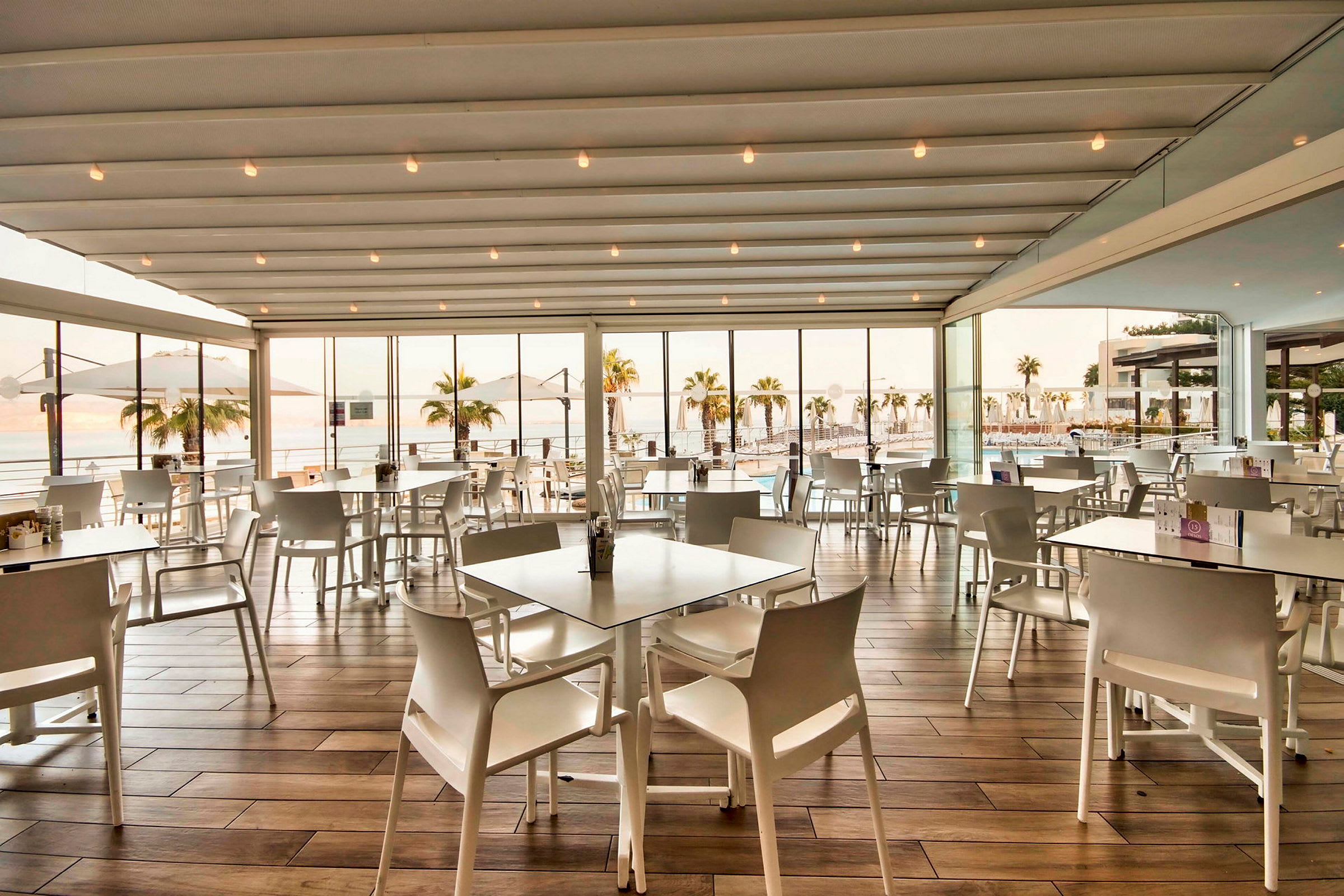 En restaurant med hvidt interiør, store vinduer og udsigt til en pool og palmer udenfor. (AI-genereret tekst)