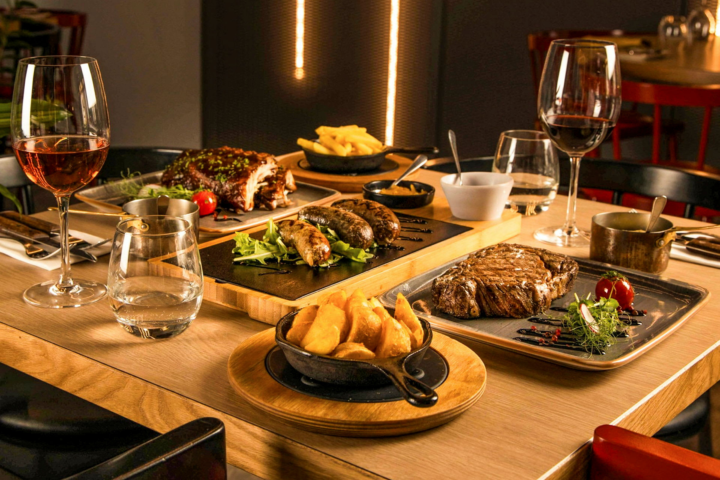 Et bord er dækket med forskellige retter, herunder bøf, pølser, spareribs og pommes frites, sammen med vinglas. (AI-genereret tekst)