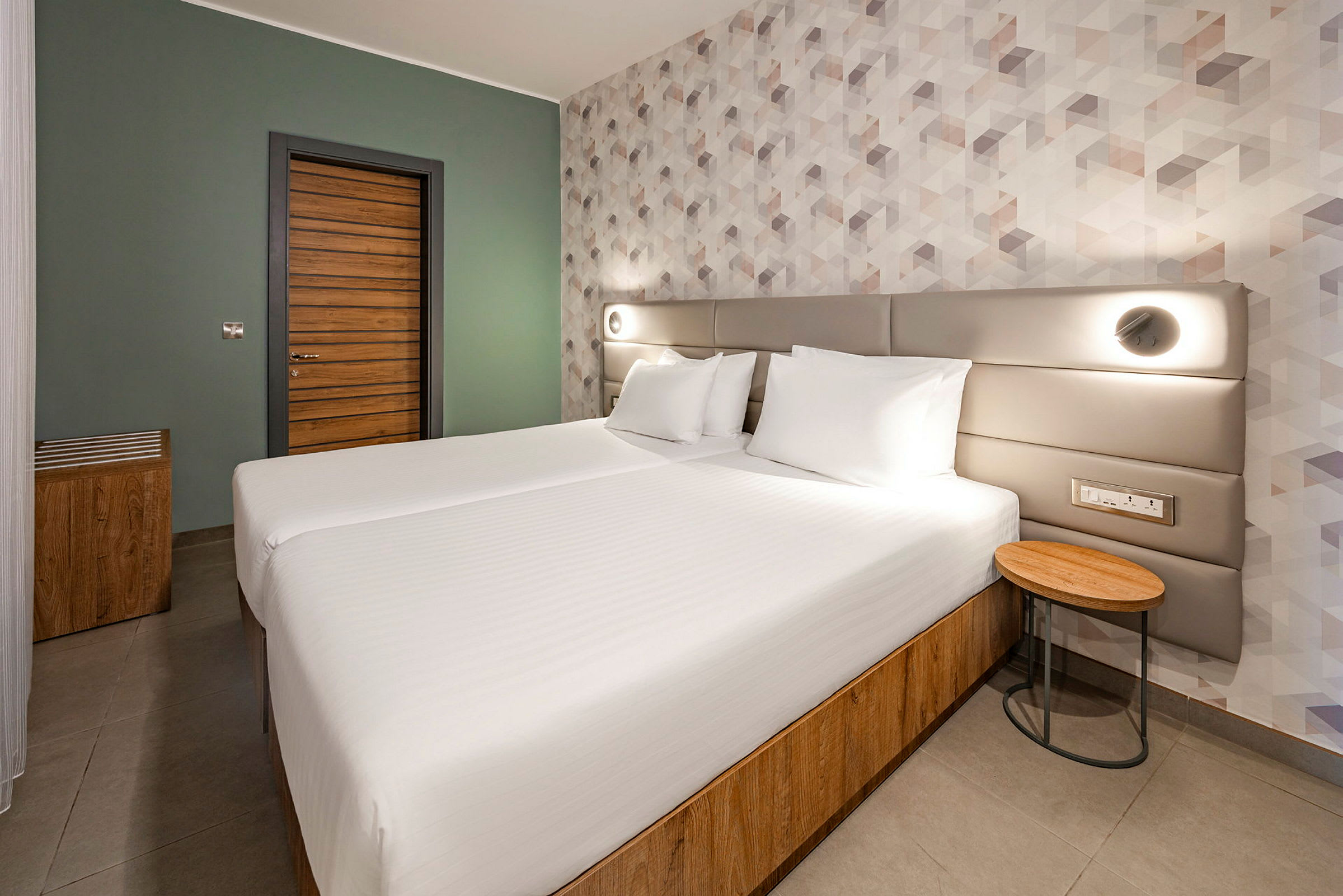 Et moderne hotelværelse med to enkeltsenge, der står sammen, en træpaneldør og geometrisk mønstrede tapeter. (AI-genereret tekst)