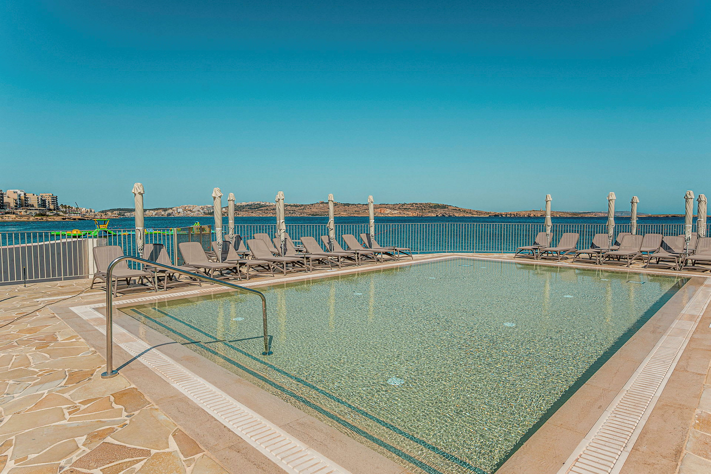 En udendørs swimmingpool med liggestole og parasoller foran en udsigt over havet og en klar himmel. (AI-genereret tekst)
