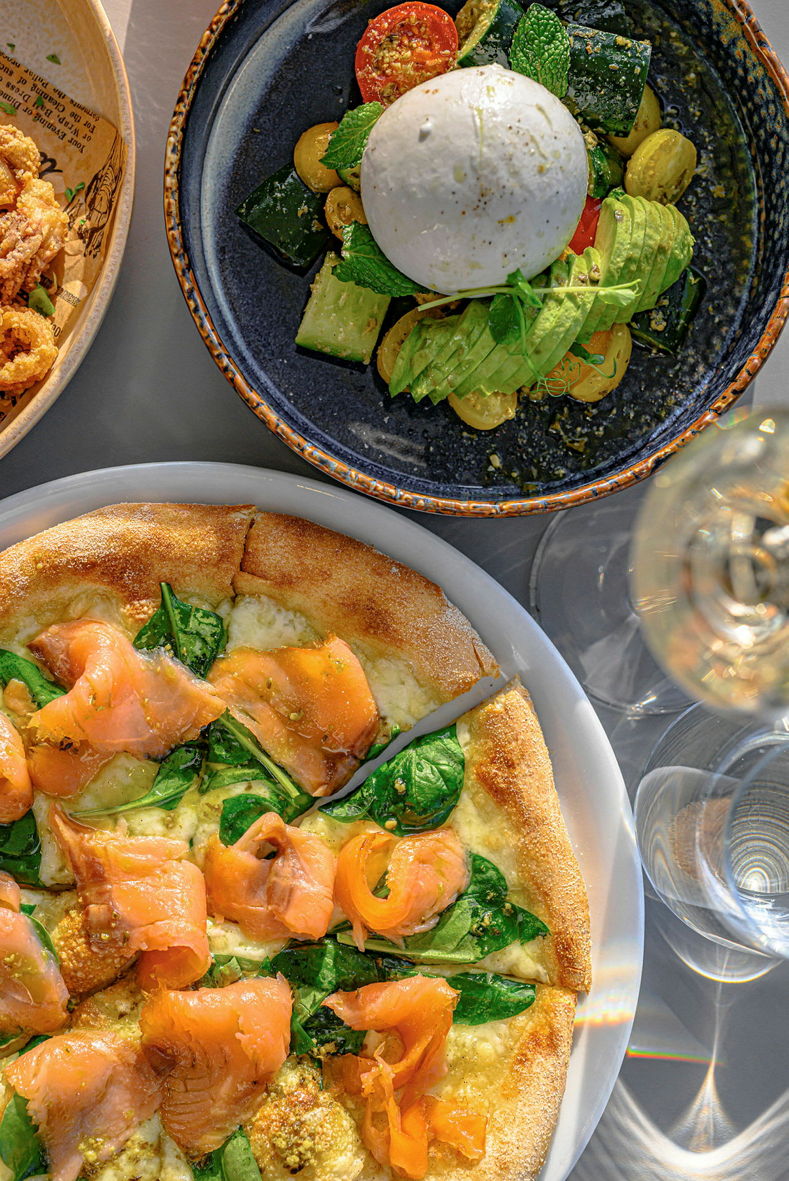 En tallerken med burrata, grøntsager og avocado, samt en pizza med laks og spinat. (AI-genereret tekst)
