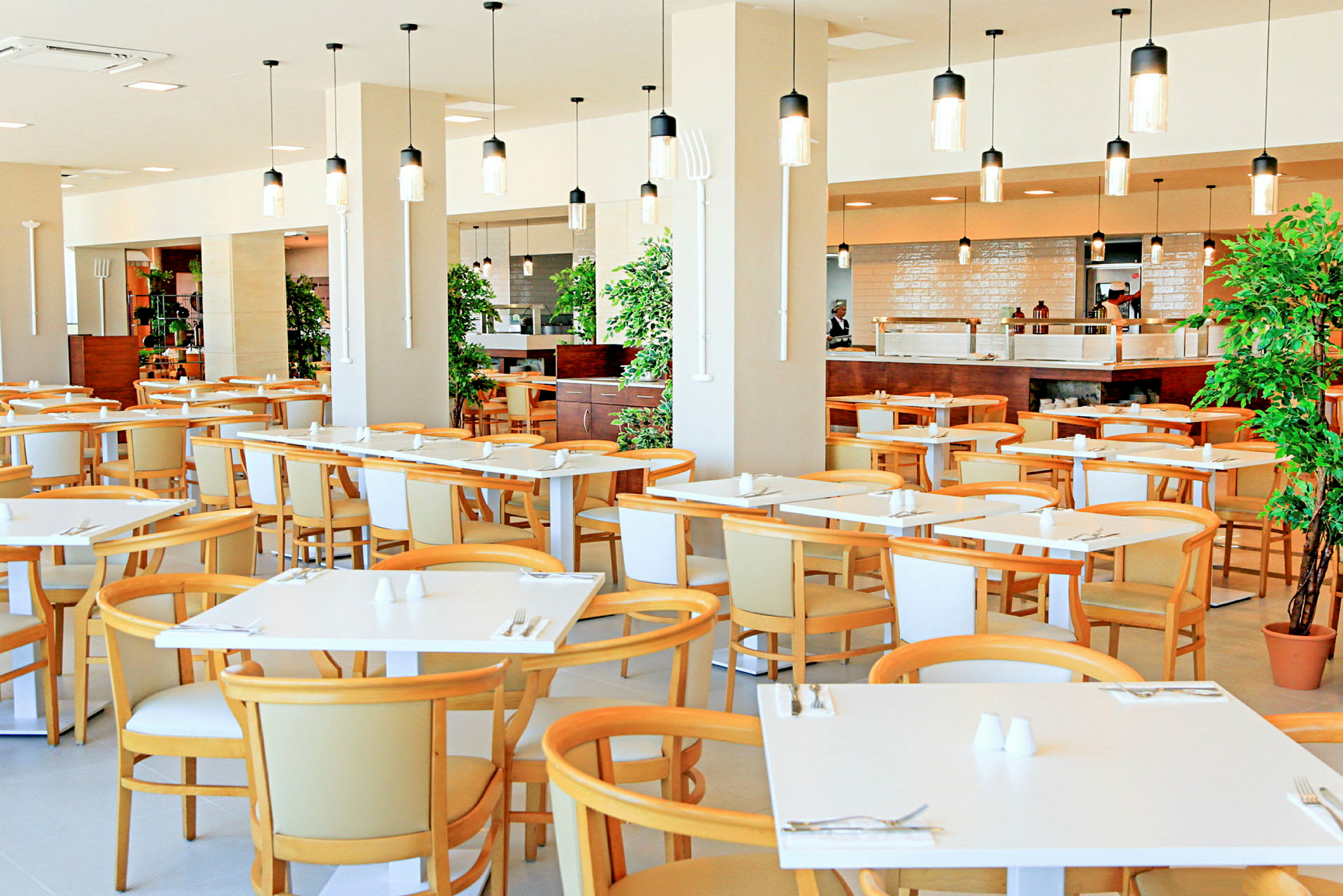 En restaurant med moderne hvide borde og lyse træstole, dekoreret med hængende lamper og planter. (AI-genereret tekst)