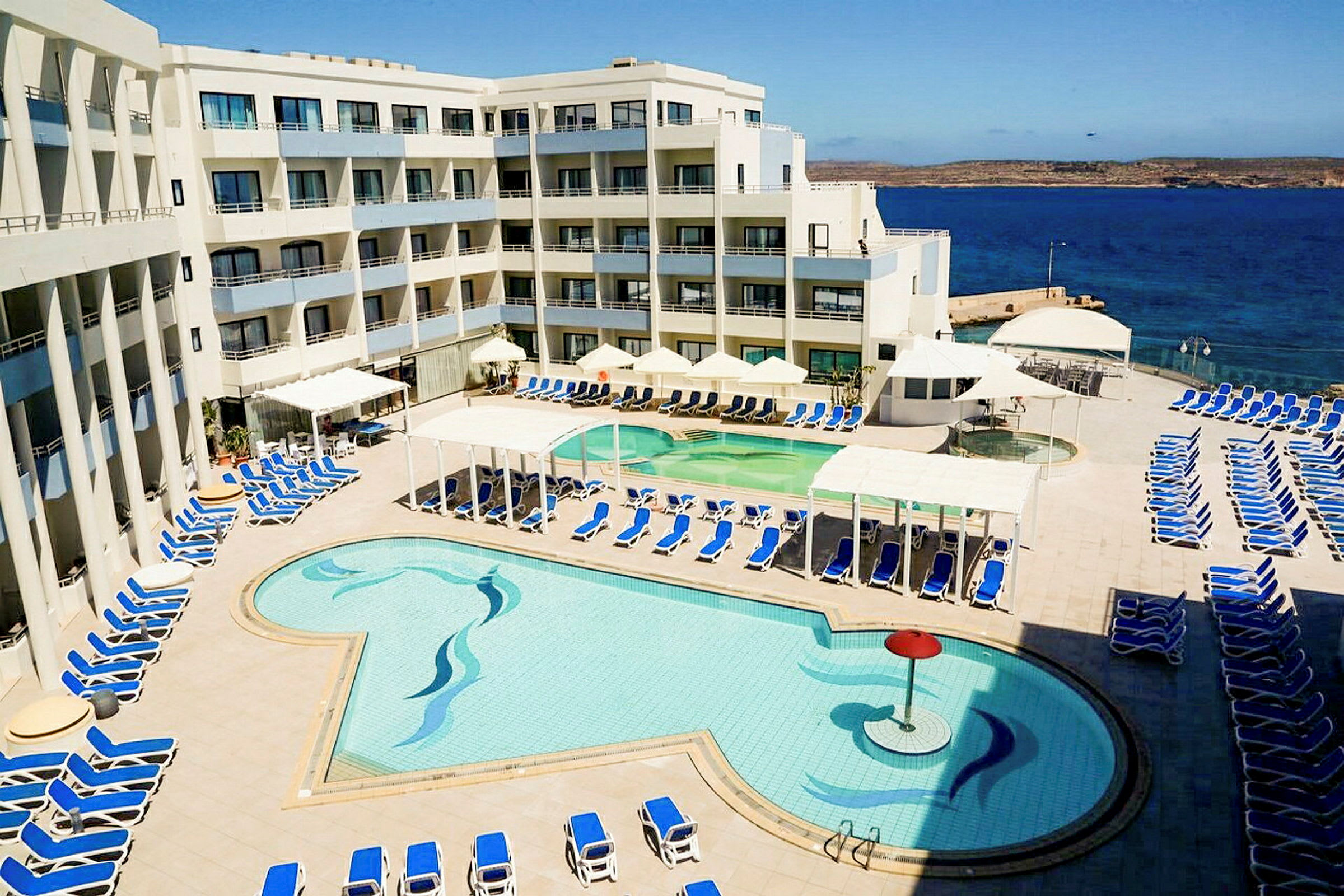 En hotelpool med liggestole og havudsigt i baggrunden. (AI-genereret tekst)