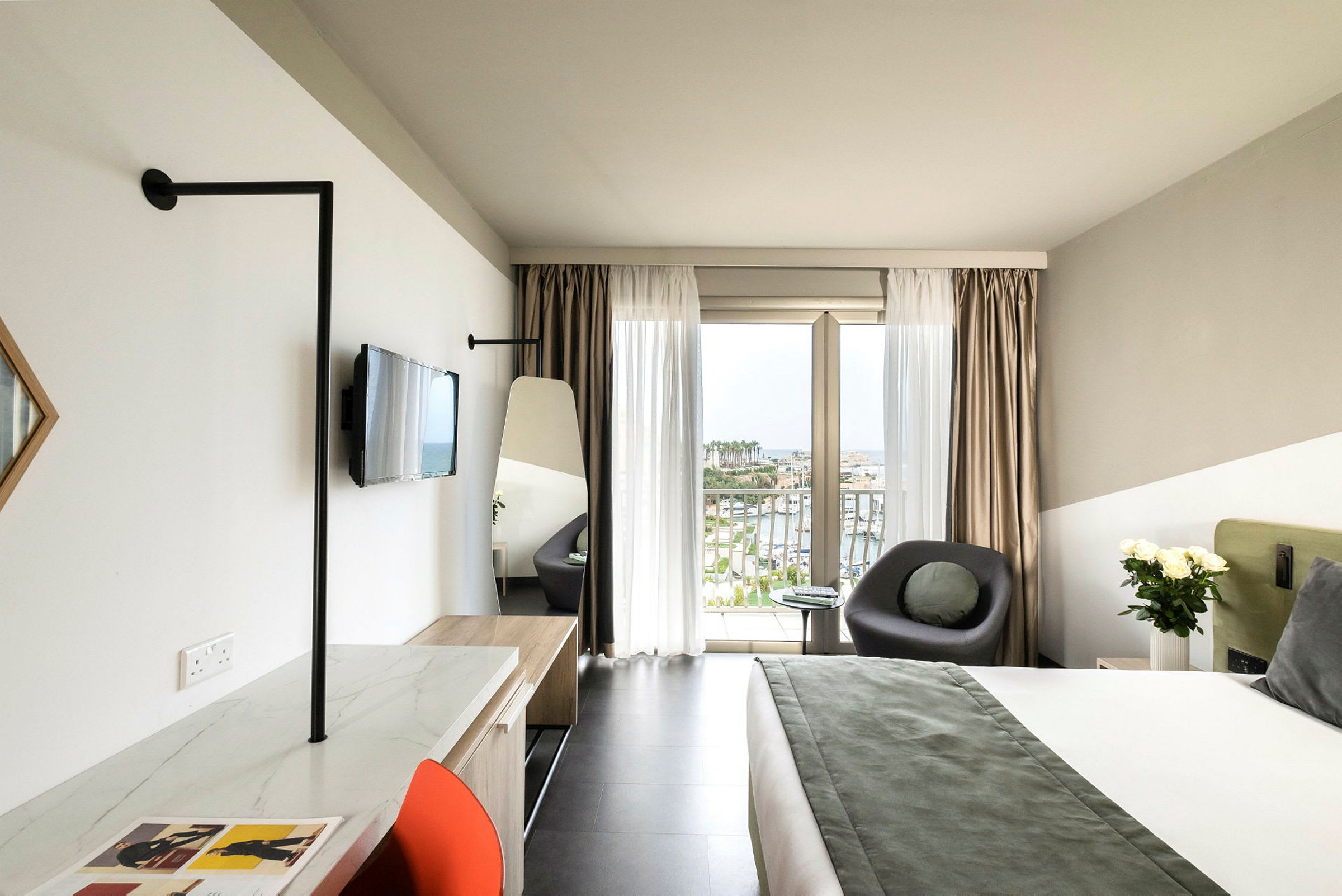 Et moderne hotelværelse med en stor seng, et siddeområde ved et vindue med udsigt over en havn, samt et skrivebord med en stol. (AI-genereret tekst)