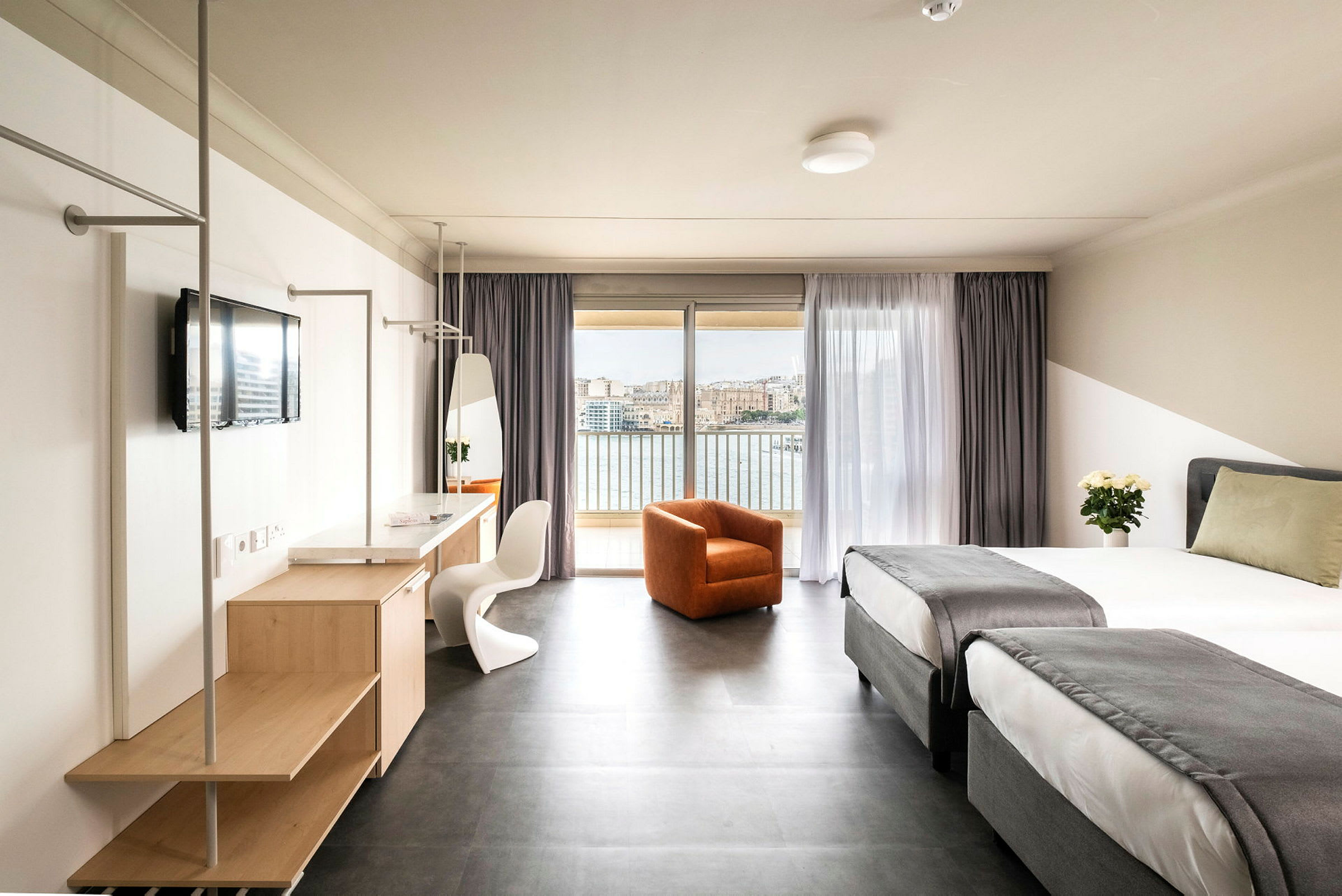 Et hotelværelse med to enkeltsenge med gråt sengetæppe, en orange lænestol, et skrivebord med spejl og tv og en balkon med udsigt over vand og en by. (AI-genereret tekst)