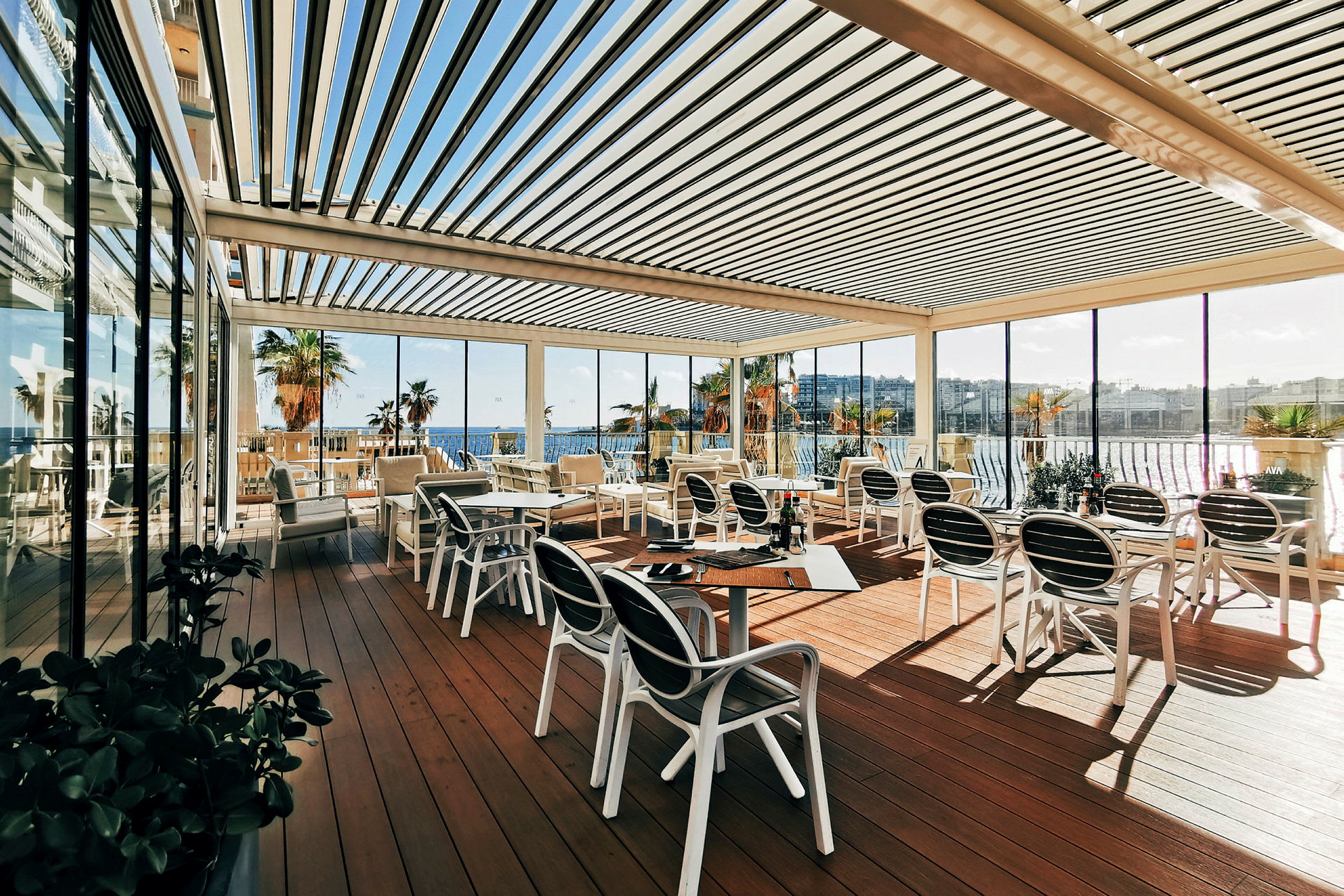 En udendørs terrasse med borde og stole, omgivet af glasvægge og med udsigt over havet og palmer. (AI-genereret tekst)