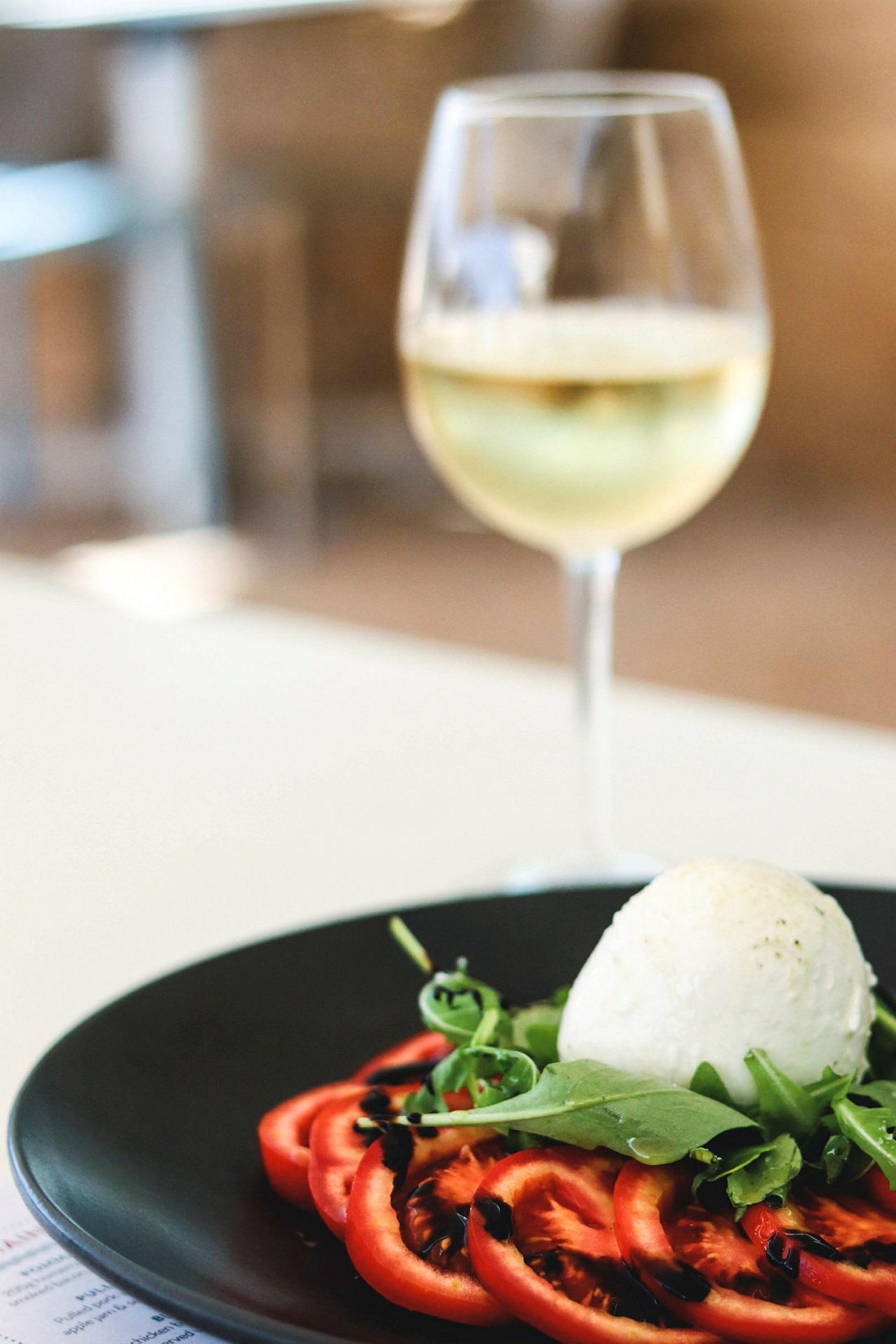 En tallerken med skiver af tomat, rucola og en kugle mozzarella, toppet med balsamico sauce. I baggrunden et glas hvidvin. (AI-genereret tekst)