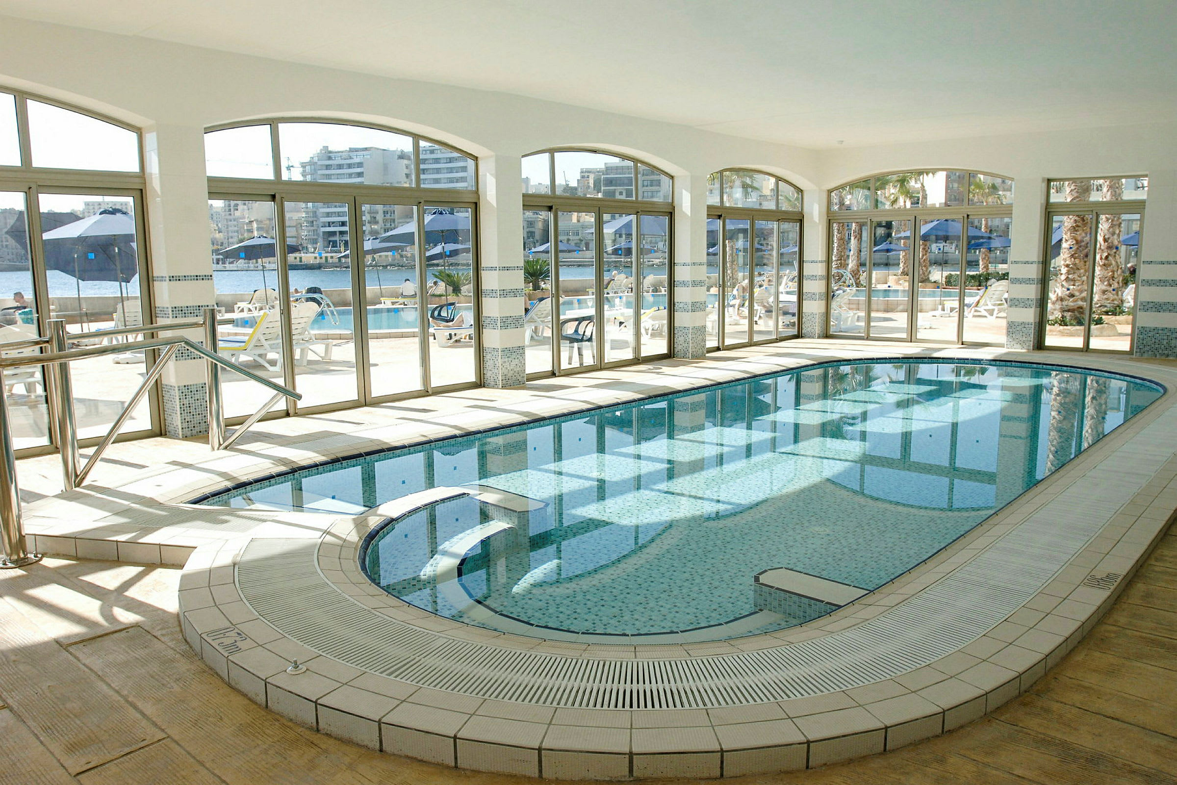En indendørs swimmingpool med glasvægge, der viser udsigten til en terrasse med siddepladser og parasoller. (AI-genereret tekst)