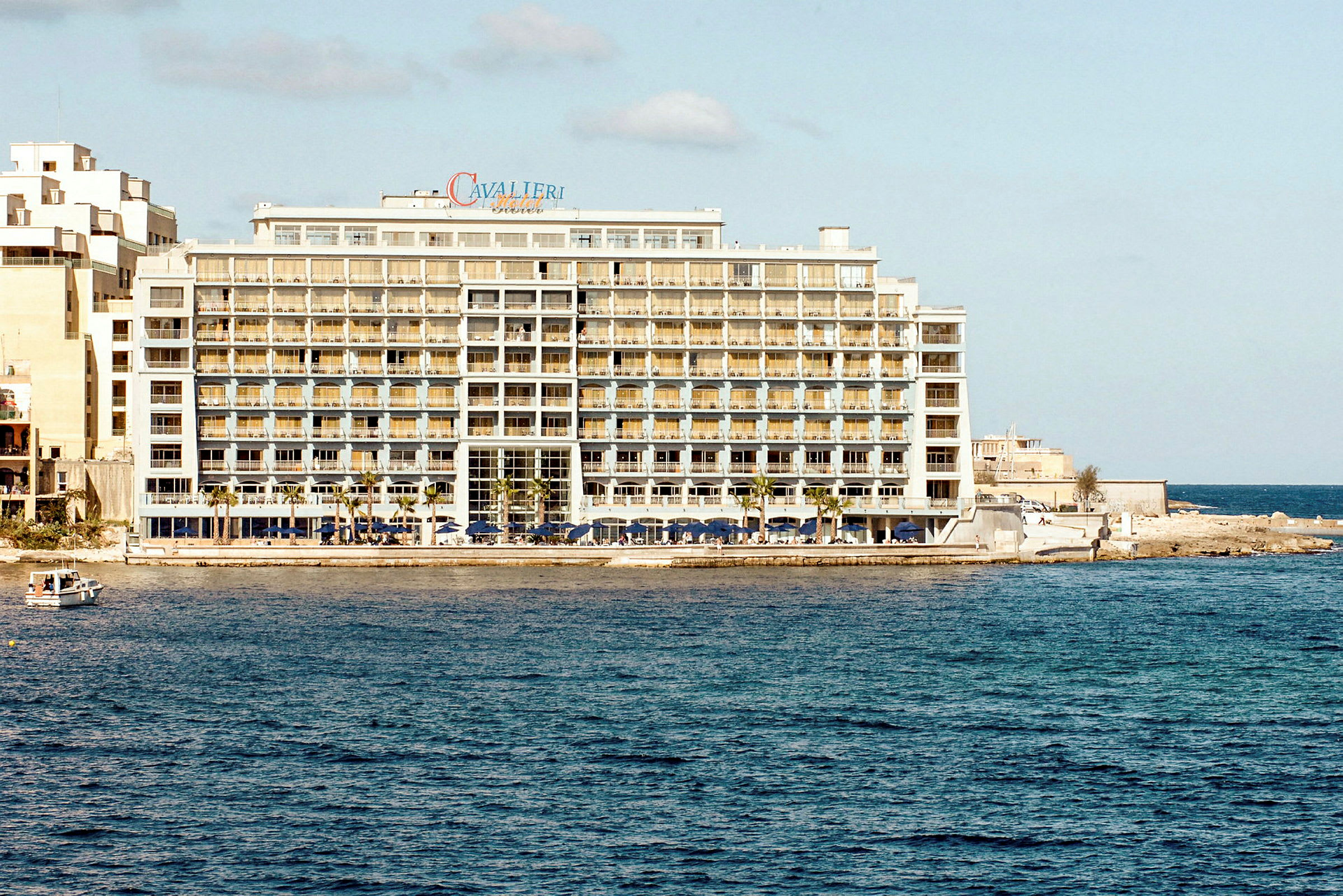 Et stort hotel ved havet med flere etager og altaner samt en lille båd i vandet tæt på hotellet. (AI-genereret tekst)