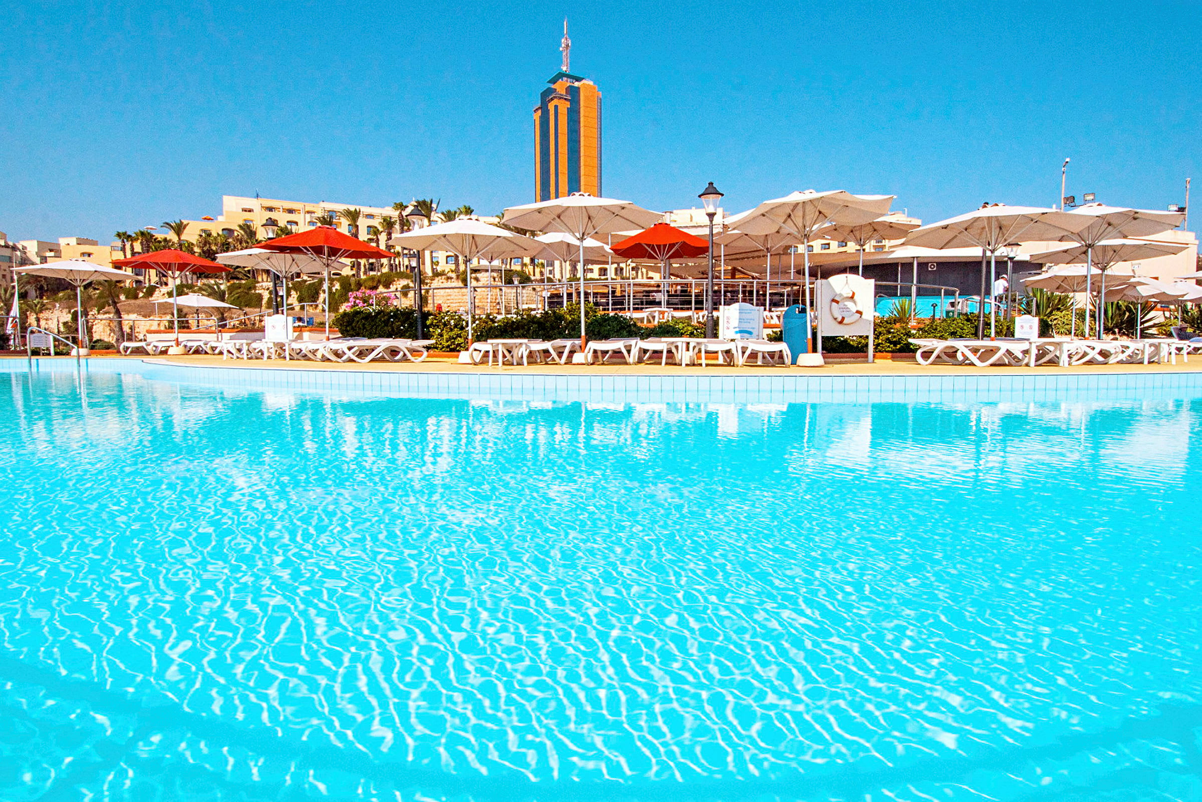 En pool med klart vand omgivet af liggestole og parasoller. I baggrunden ses bygninger og en høj bygning. (AI-genereret tekst)