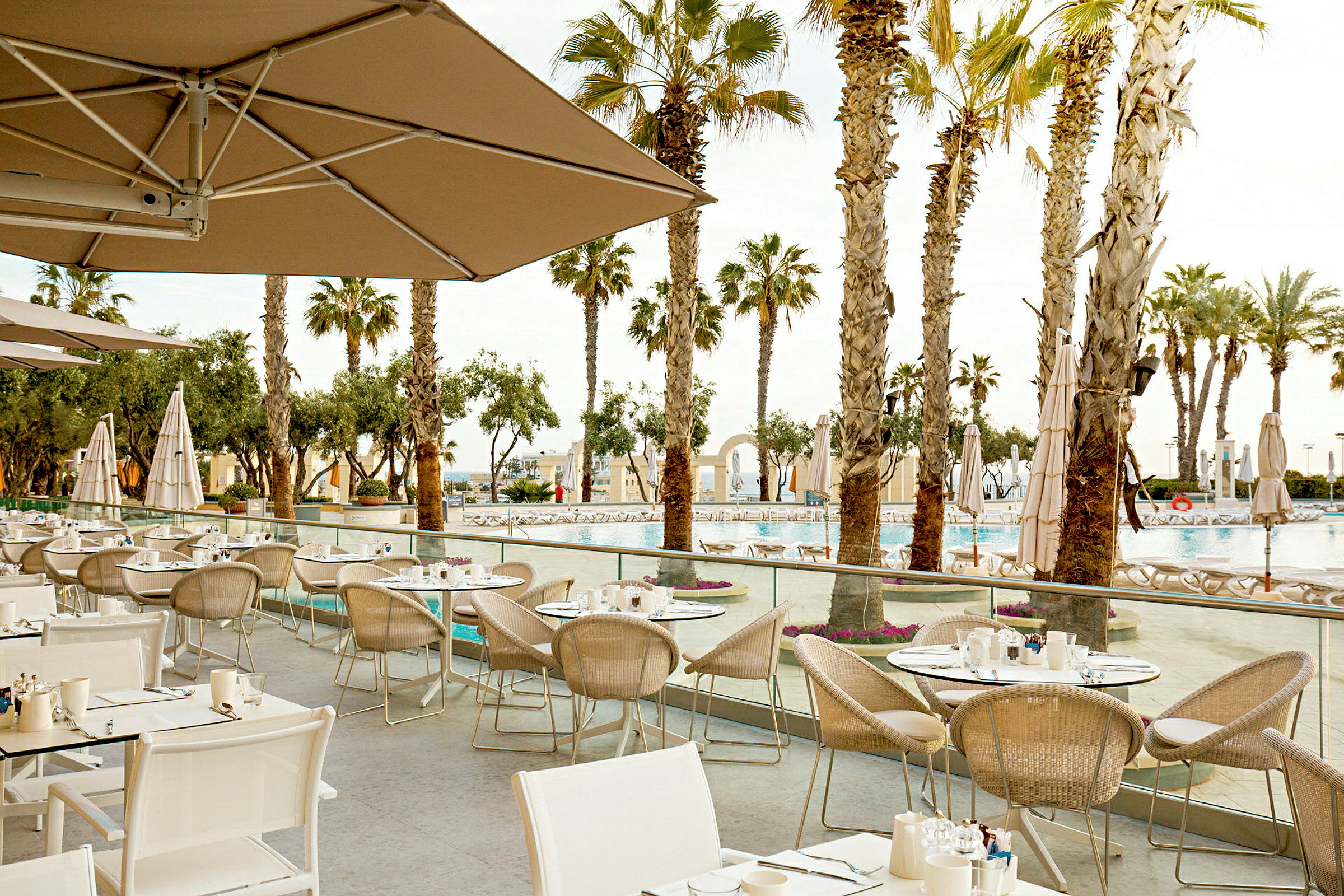 En udendørs restaurant med flere opstillede borde og stole, omgivet af palmer og med udsigt over en pool. (AI-genereret tekst)