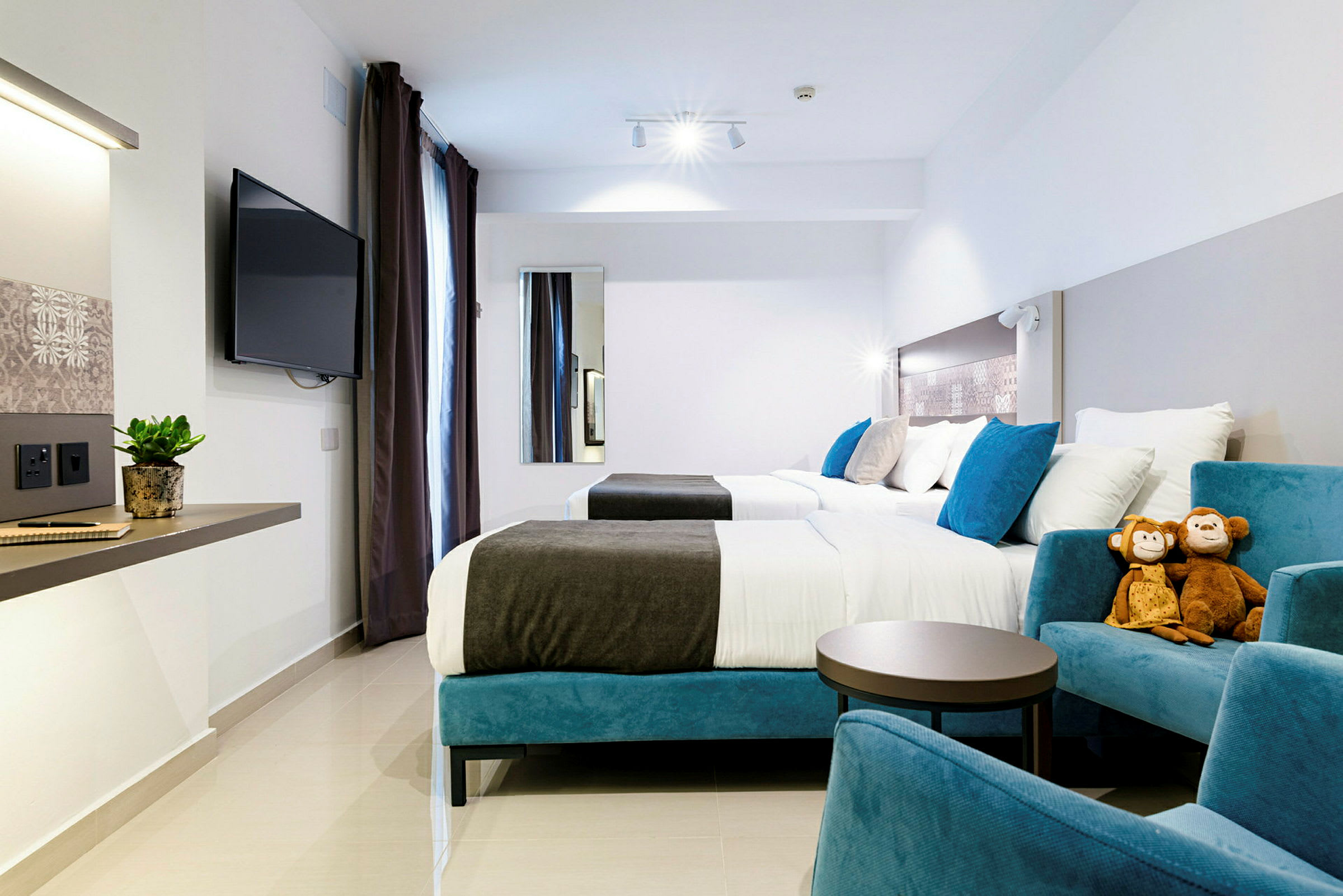 Et moderne hotelværelse med to senge, siddepladser, et vægmonteret TV og to plysdyr på en stol. (AI-genereret tekst)