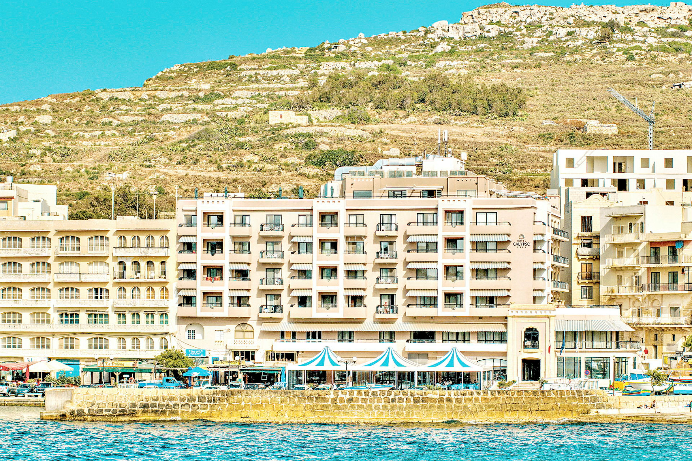 Hotellets facade ved strandpromenaden med kuperet terræn i baggrunden. (AI-genereret tekst)